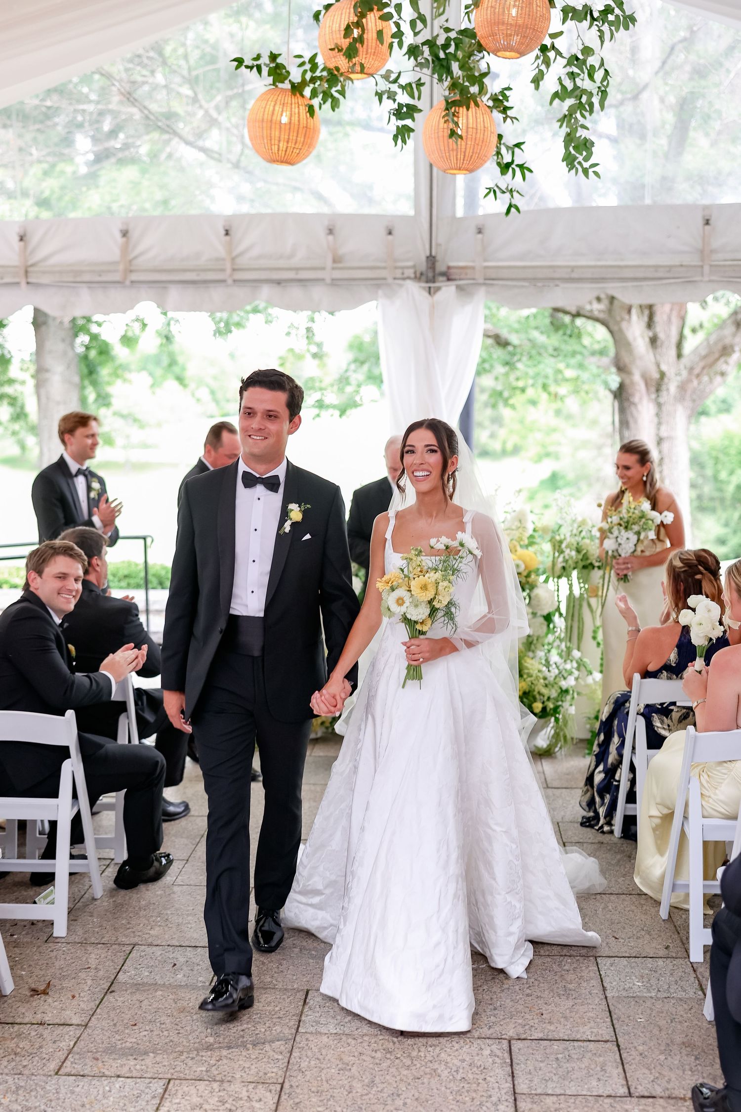 Bride and groom walk down the aisle hand in hand under a white tent adorned with lanterns and greenery.