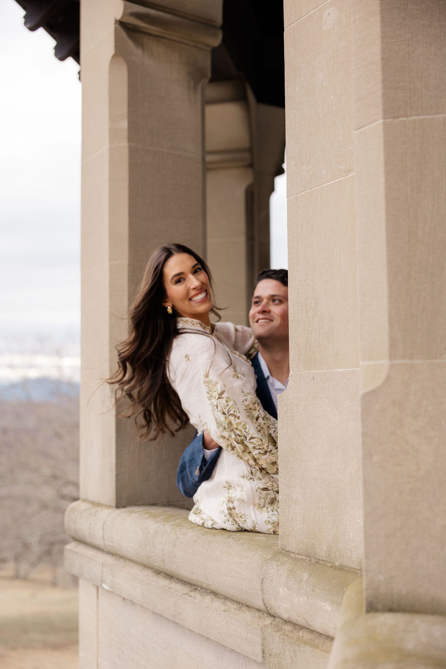 A couple shares a joyful moment while posing together between stone pillars on a scenic overlook.