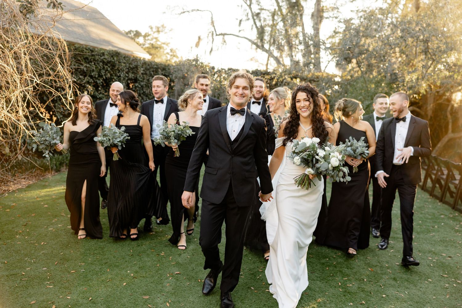 Wedding party walks down outdoor path with bride in white dress and attendants in black attire.