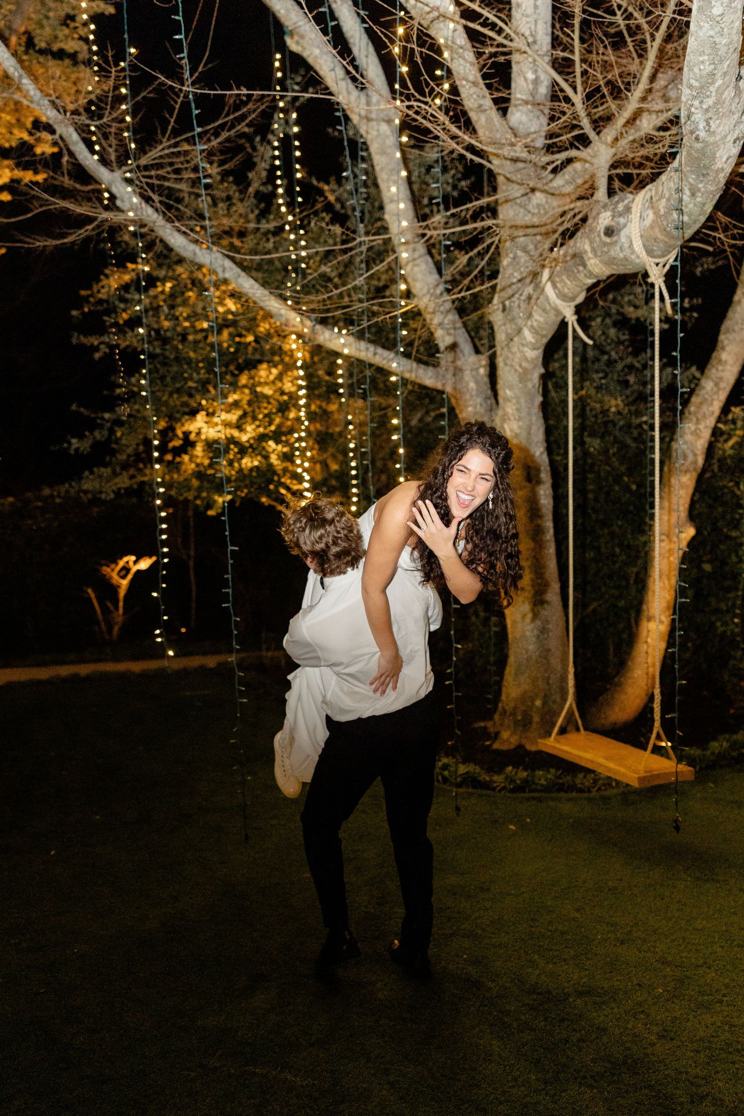 A couple dances under twinkling lights strung in trees during an evening wedding celebration.