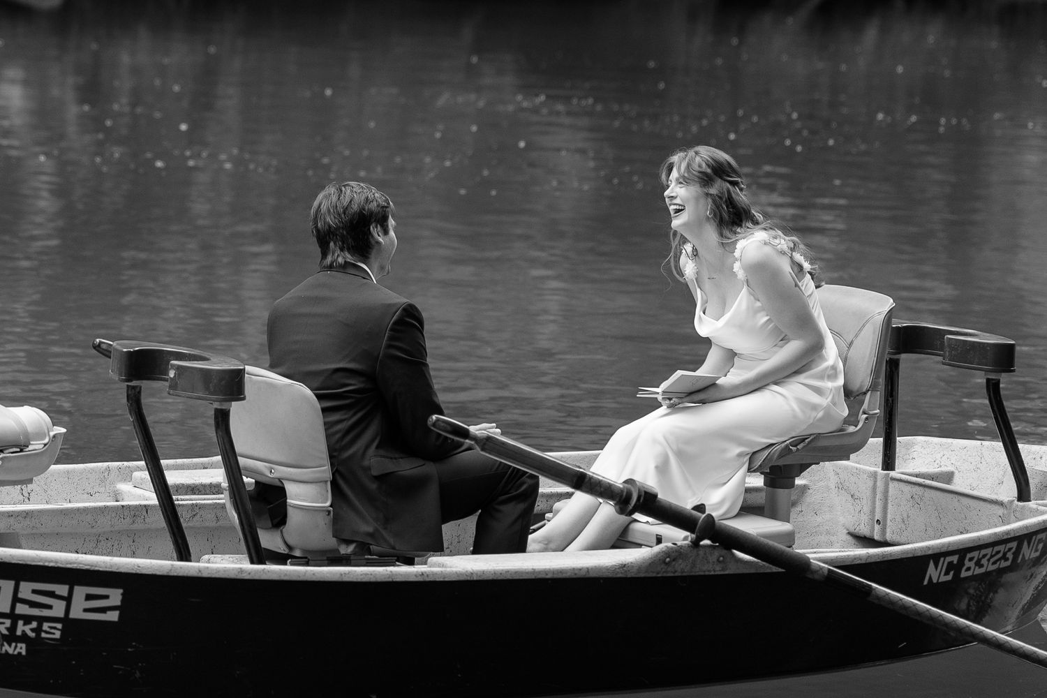 Black and white photo of a bride and groom sharing a romantic moment on a rowboat on a serene lake.