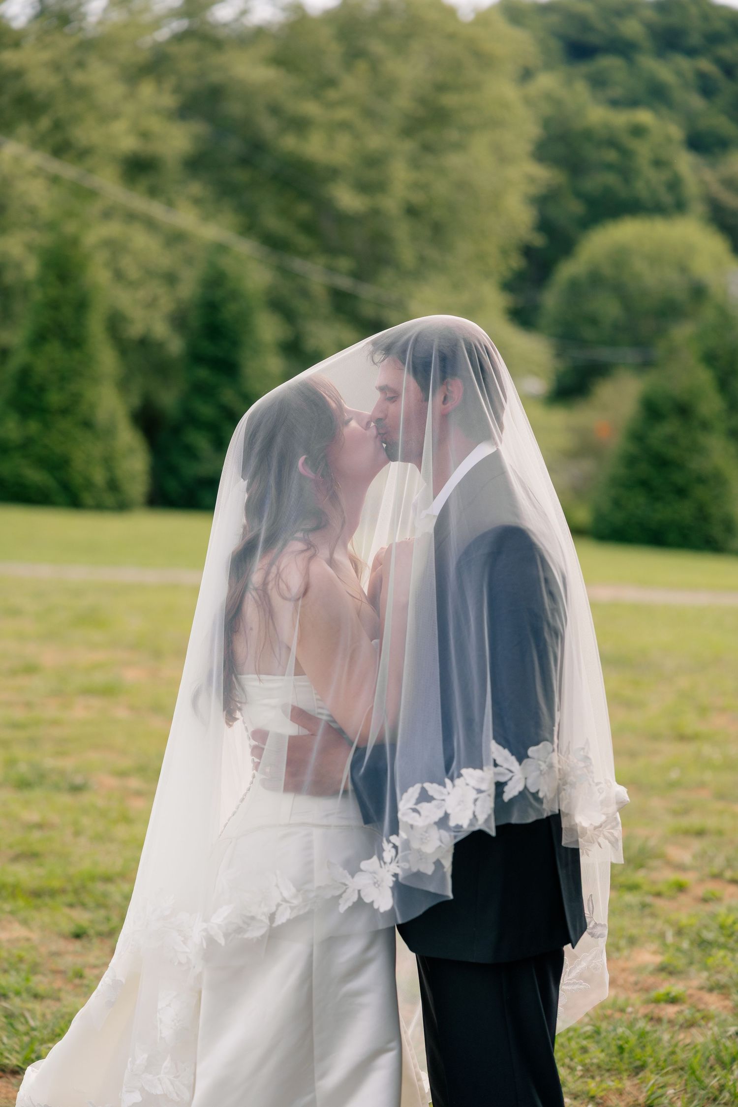 Bride and groom share a kiss hidden beneath a flowing lace-trimmed veil outdoors.
