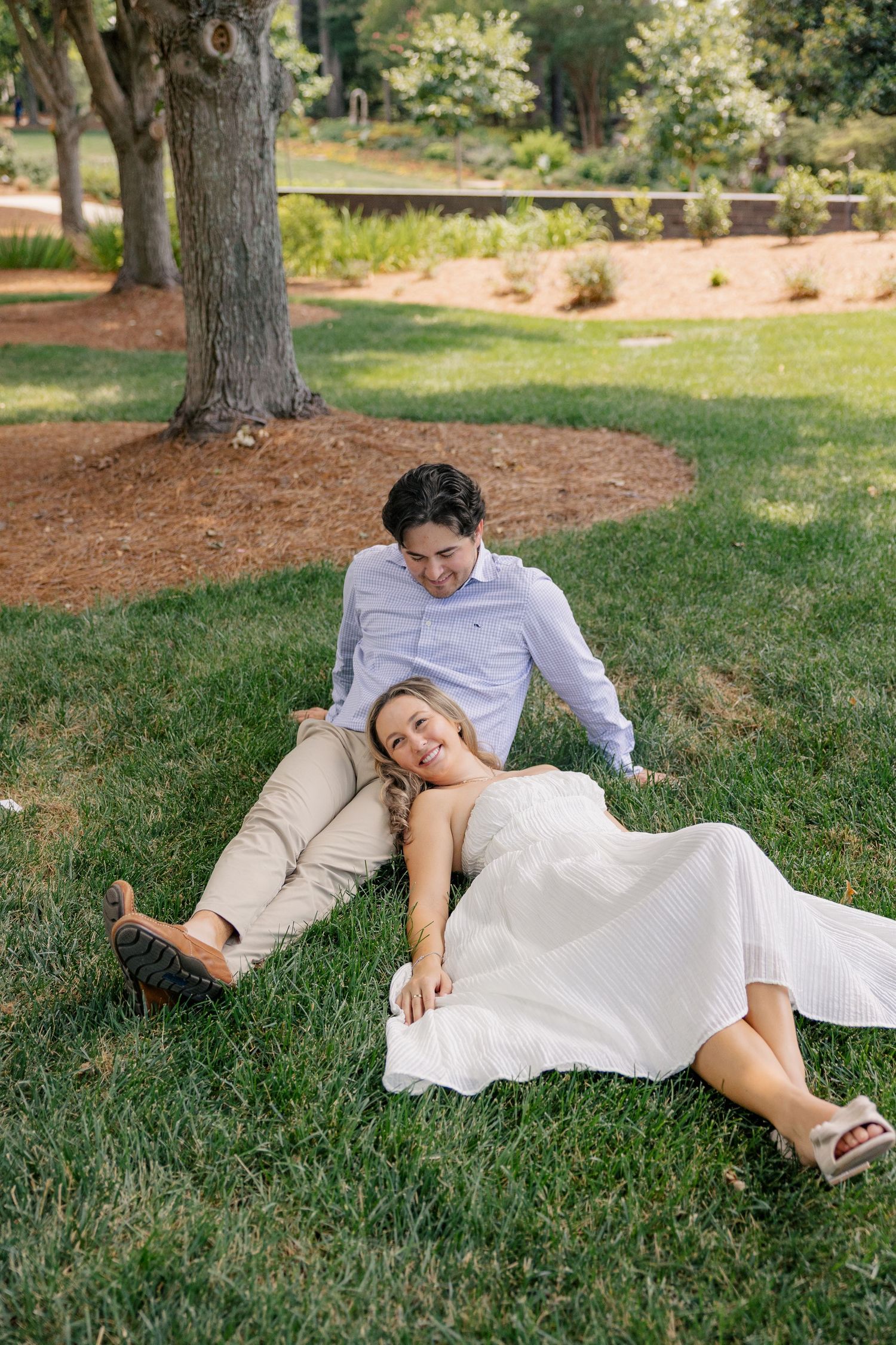 Couple relaxing on green grass under tree, woman in white dress laughing while resting on mans lap.