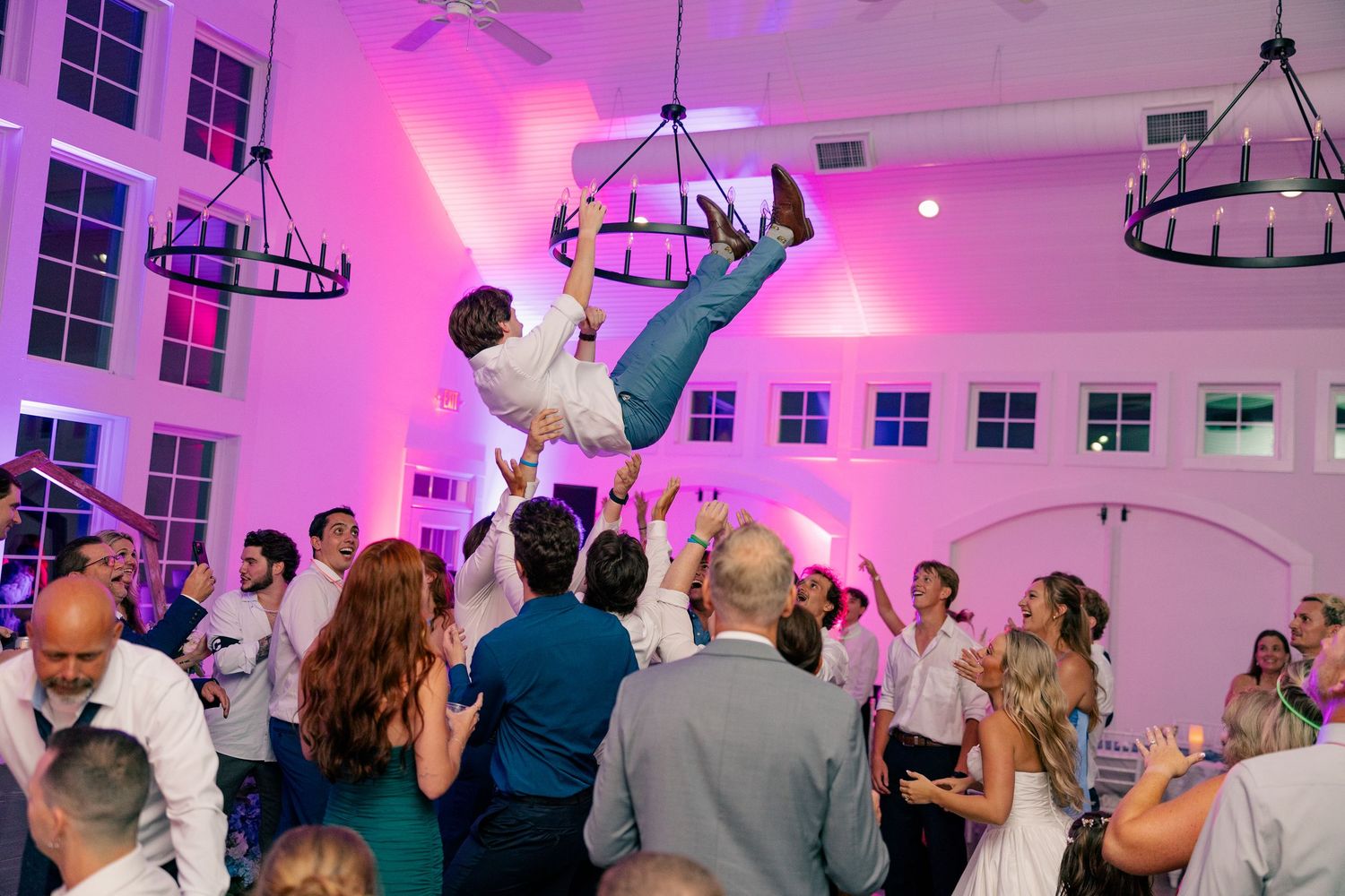 Groom lifted by wedding guests on a pink-lit dance floor during a lively wedding reception celebration.