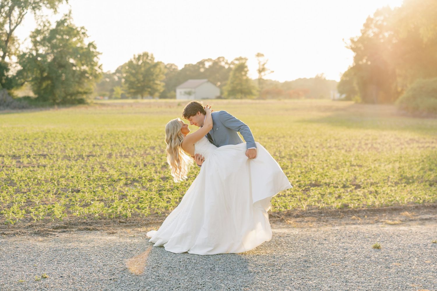 Bride and groom share romantic dip kiss on gravel path surrounded by golden sunset farm scenery.