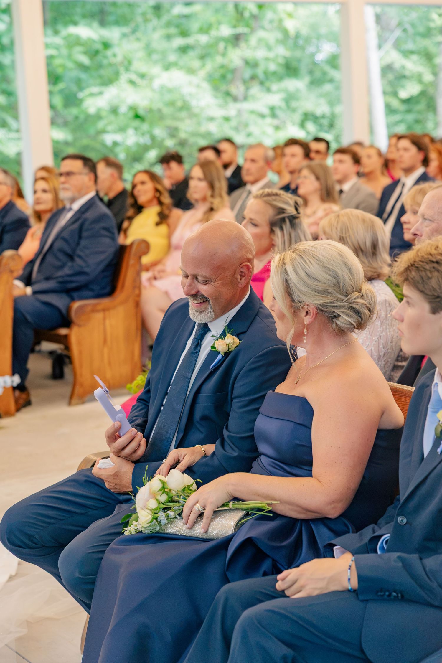 Wedding guests in navy attire smile and laugh during an indoor ceremony with large windows.