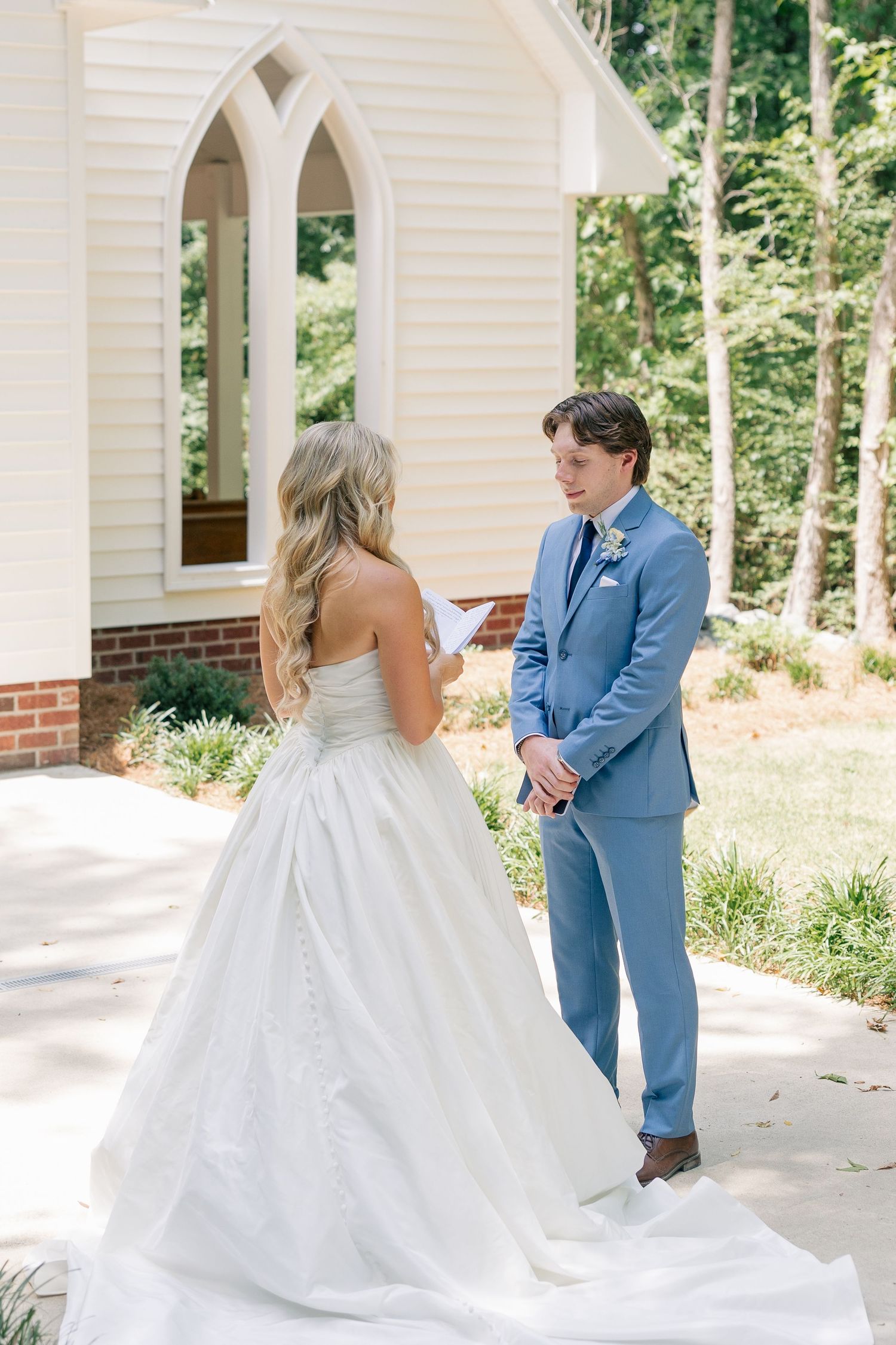 Groom reads vows as bride stands before a white chapel surrounded by trees.