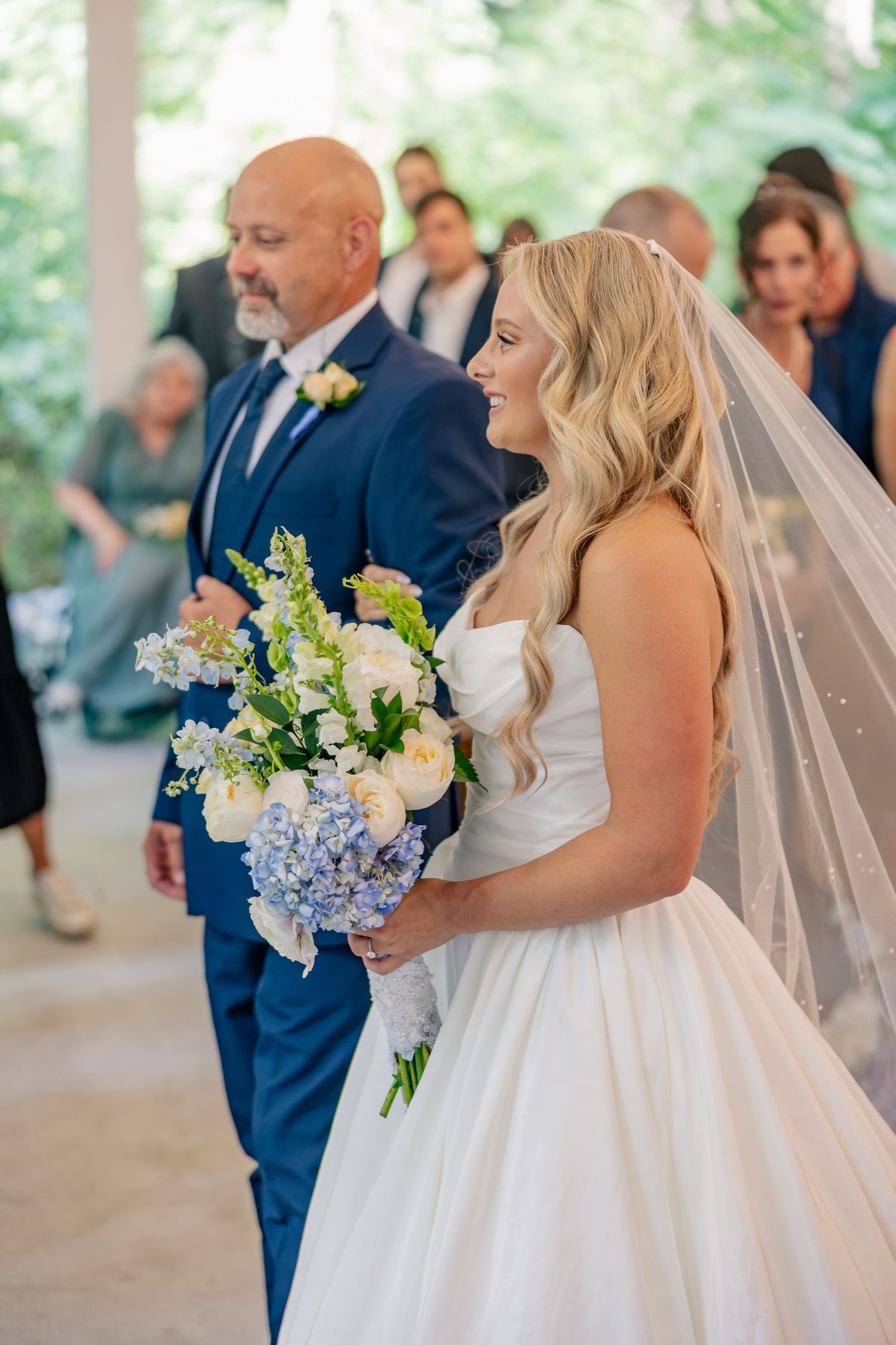 Smiling bride in white strapless gown holding blue and white bouquet walking down the aisle.