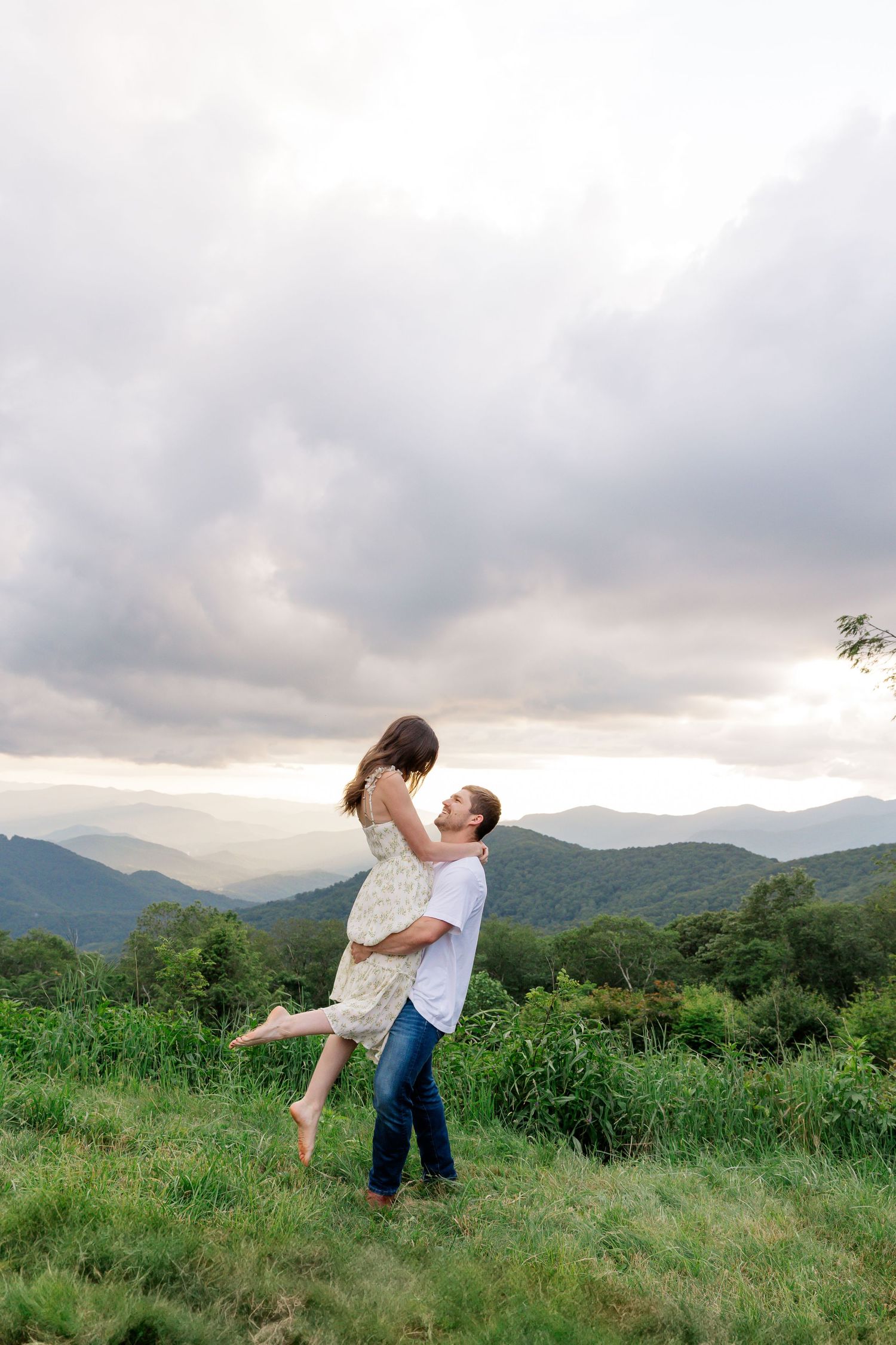 A romantic couple dances together in a grassy meadow with misty mountains in the background at sunset.