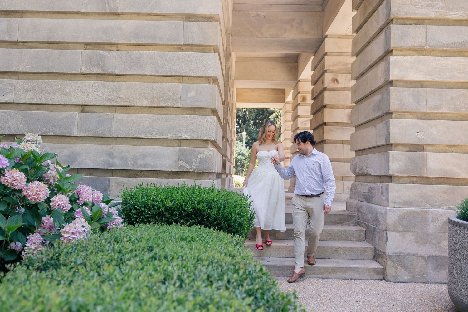 Couple in elegant attire walking along stone steps near blooming hydrangeas at a classic building.