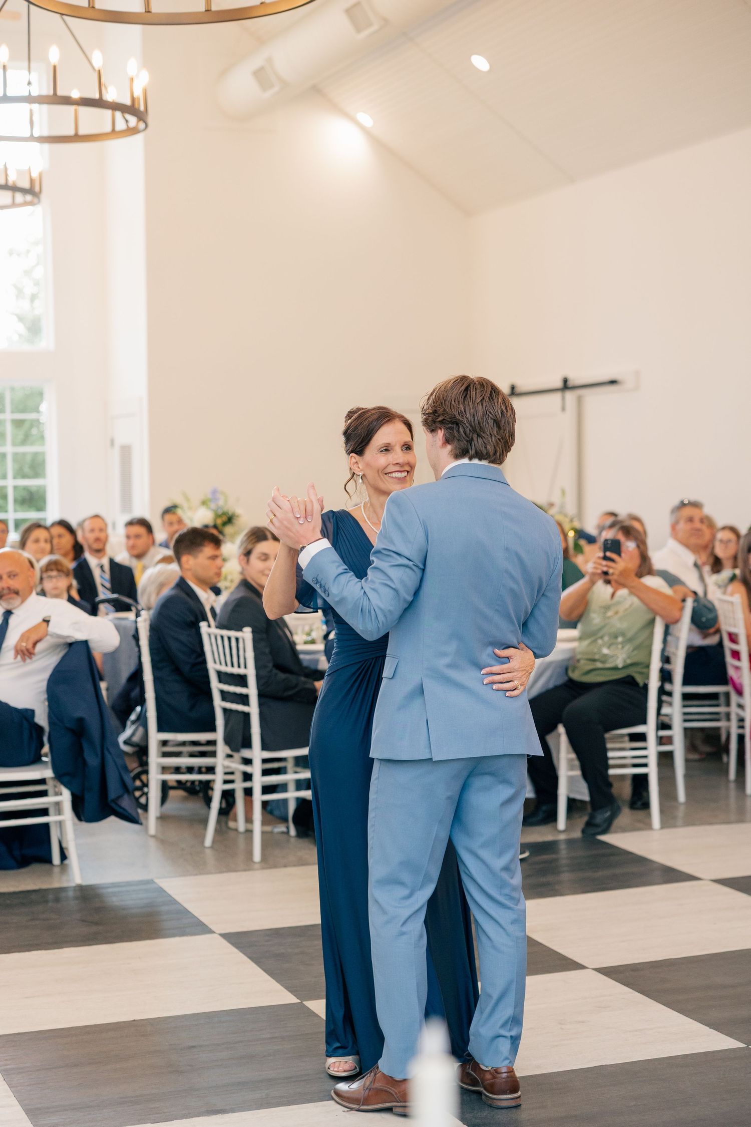 Groom in light blue suit dances with smiling woman in navy dress at wedding reception.