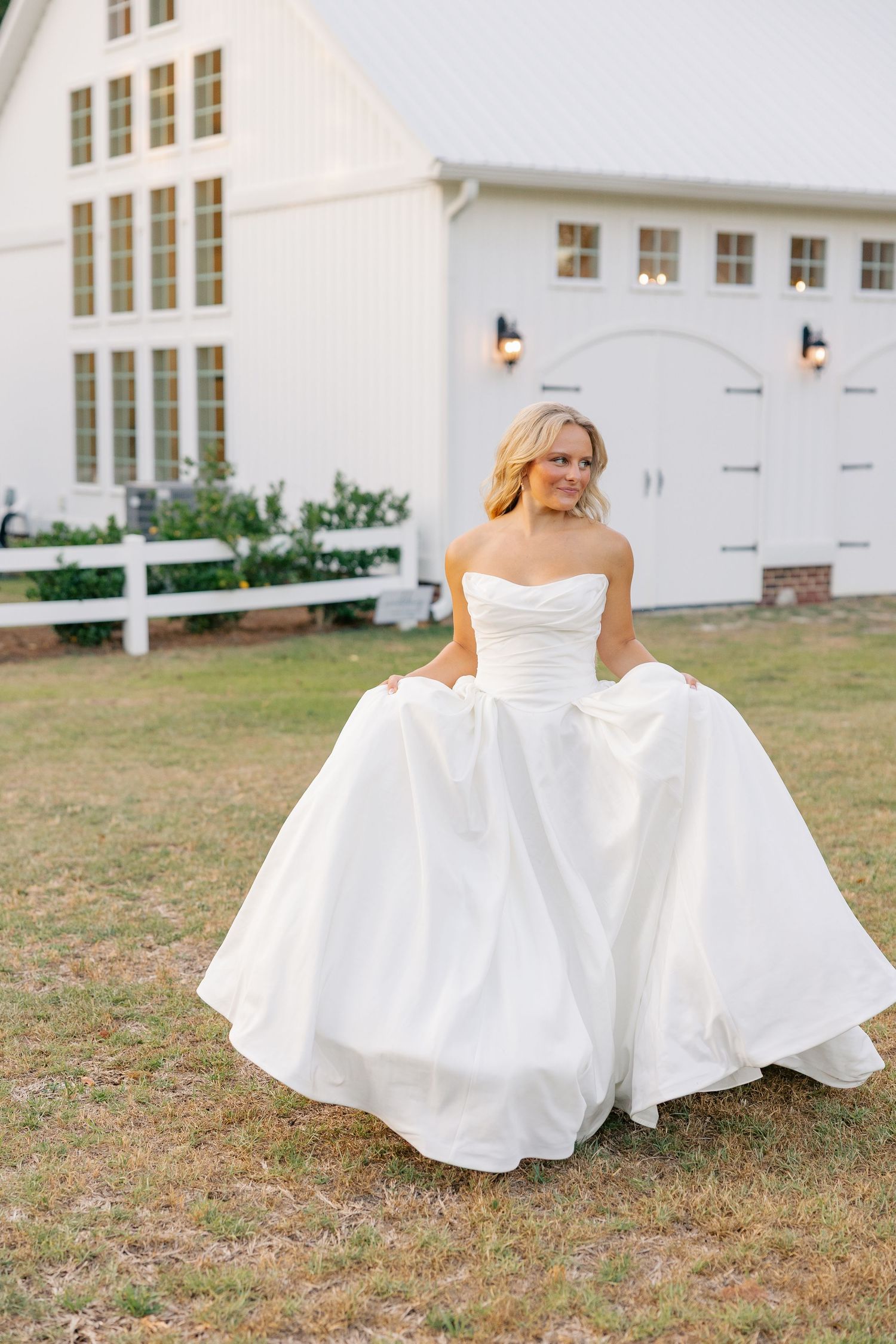 Blonde bride twirls in strapless white ball gown outside a white barn wedding venue at dusk.
