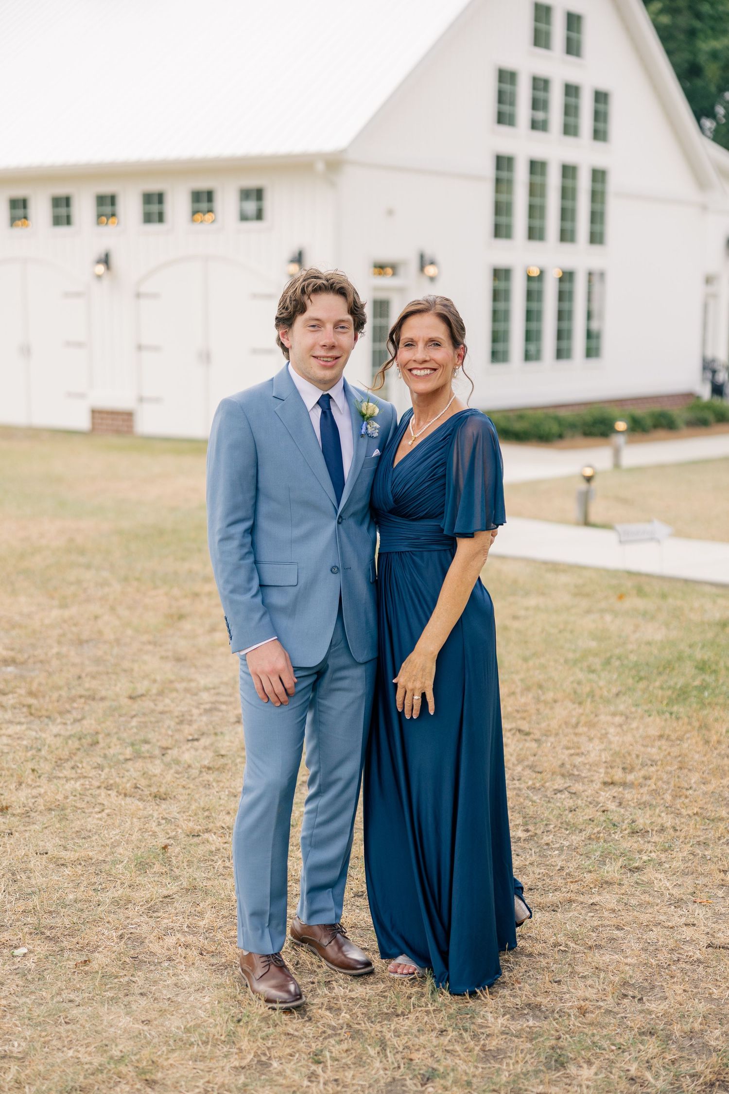 Groom in light blue suit posing with woman in navy dress outside white barn wedding venue.