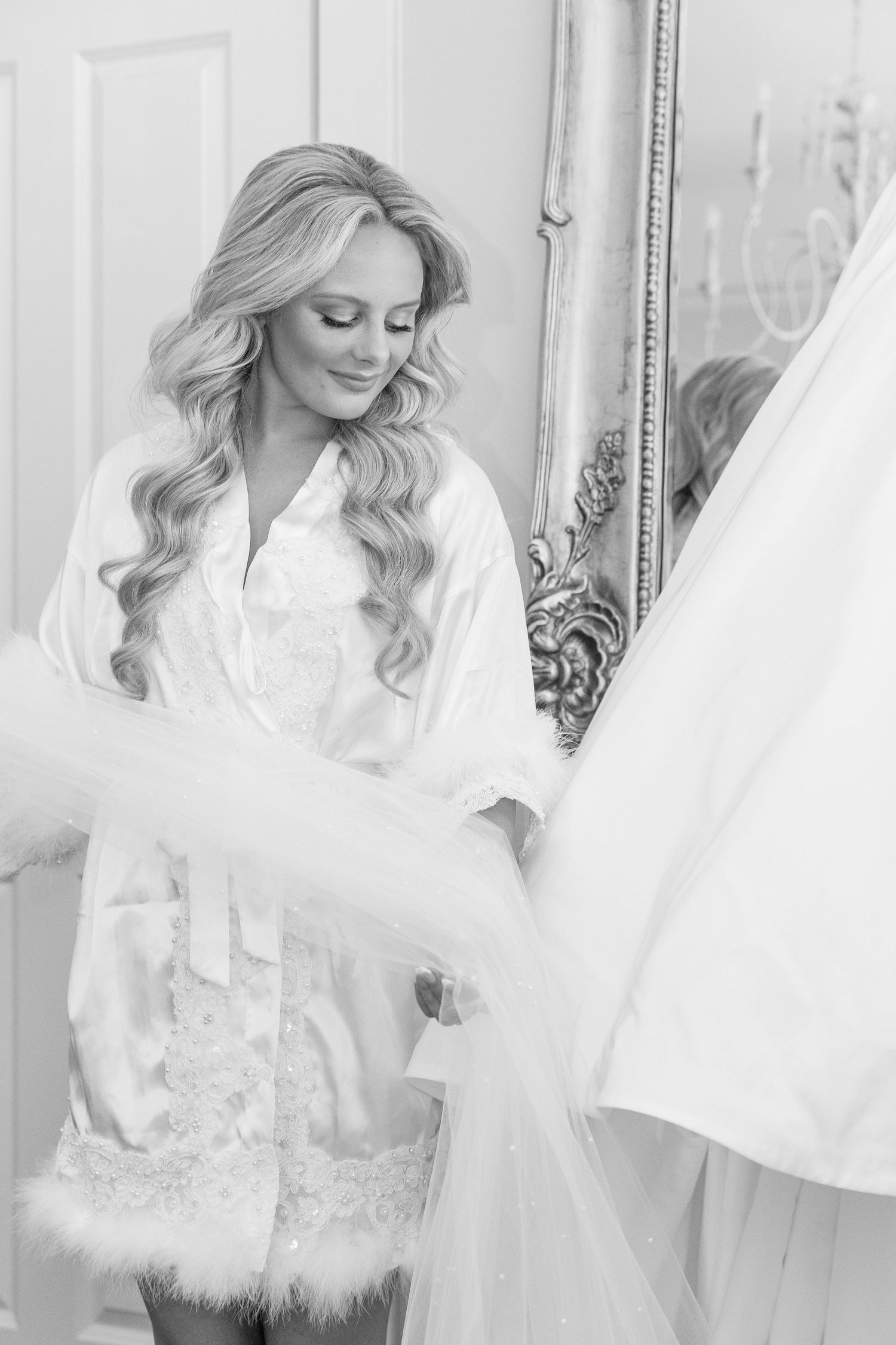 Bride in white lace robe smiling while holding her wedding dress in front of an ornate mirror.