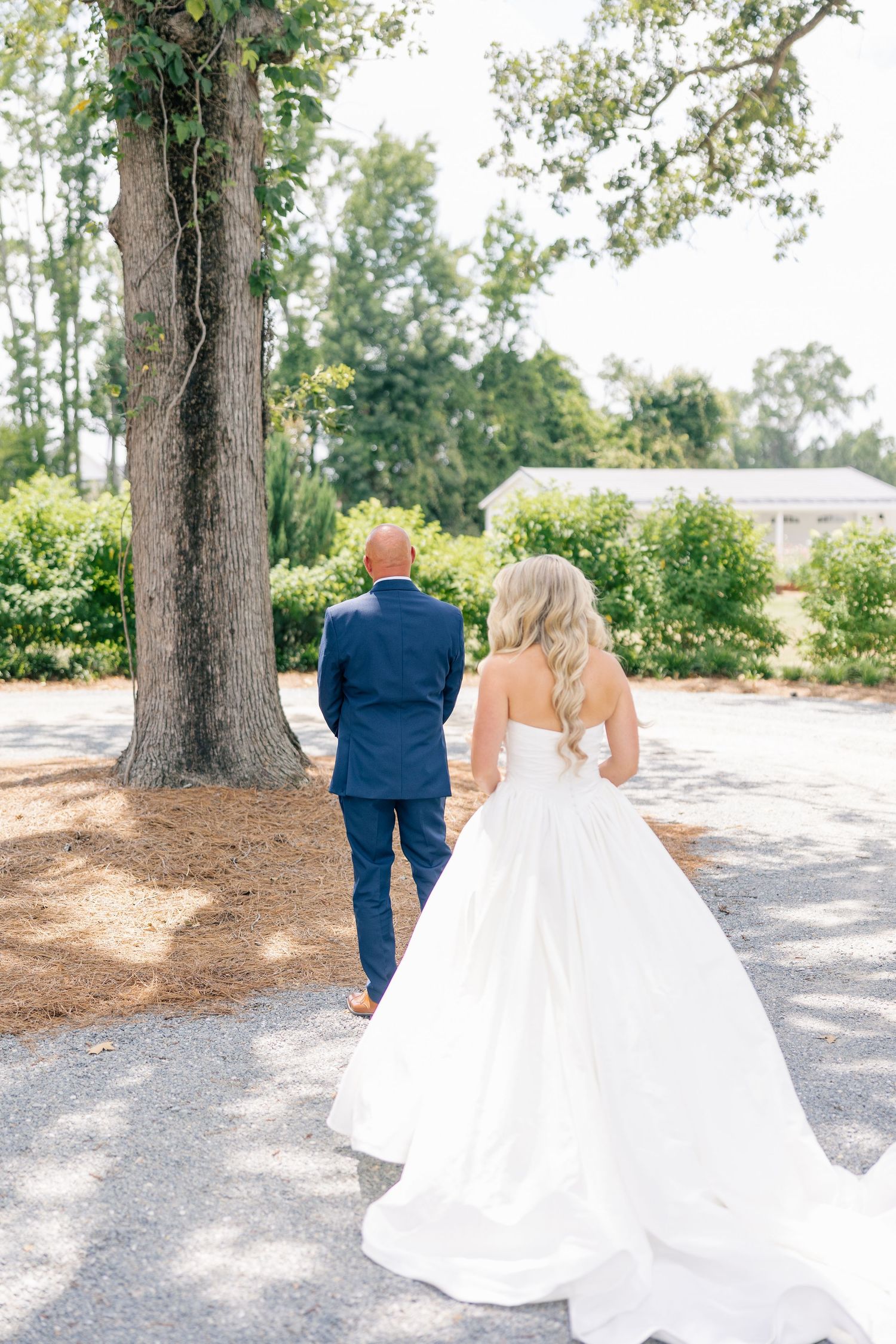 Bride in strapless white ball gown walks with a man in a navy suit toward a tree-lined path.