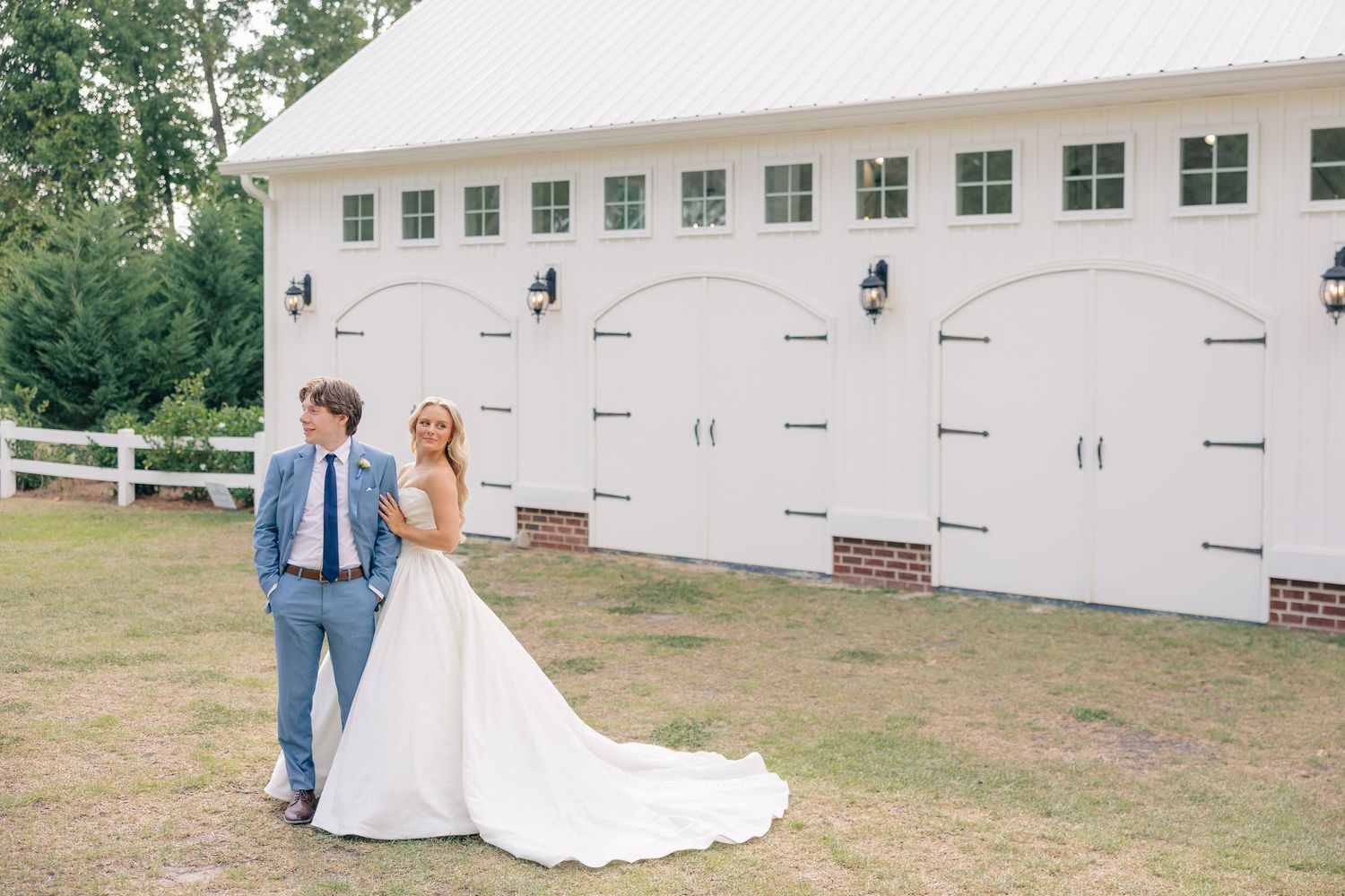 Bride and groom stand together on lawn in front of white barn wedding venue.