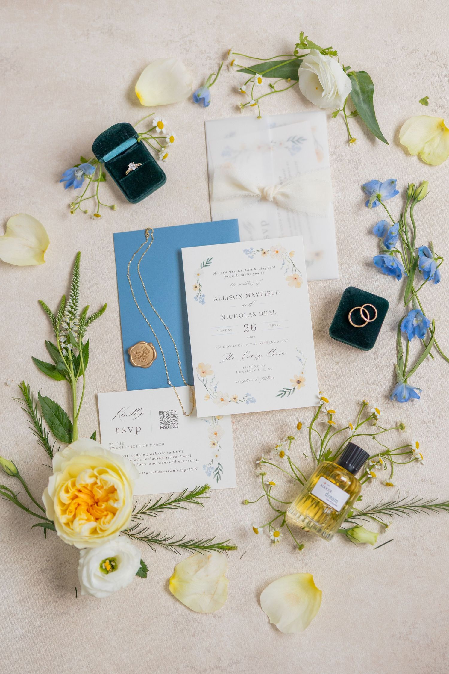 Elegant wedding flat lay with blue envelope, floral invitation, gold rings, perfume, and fresh flowers on neutral background.