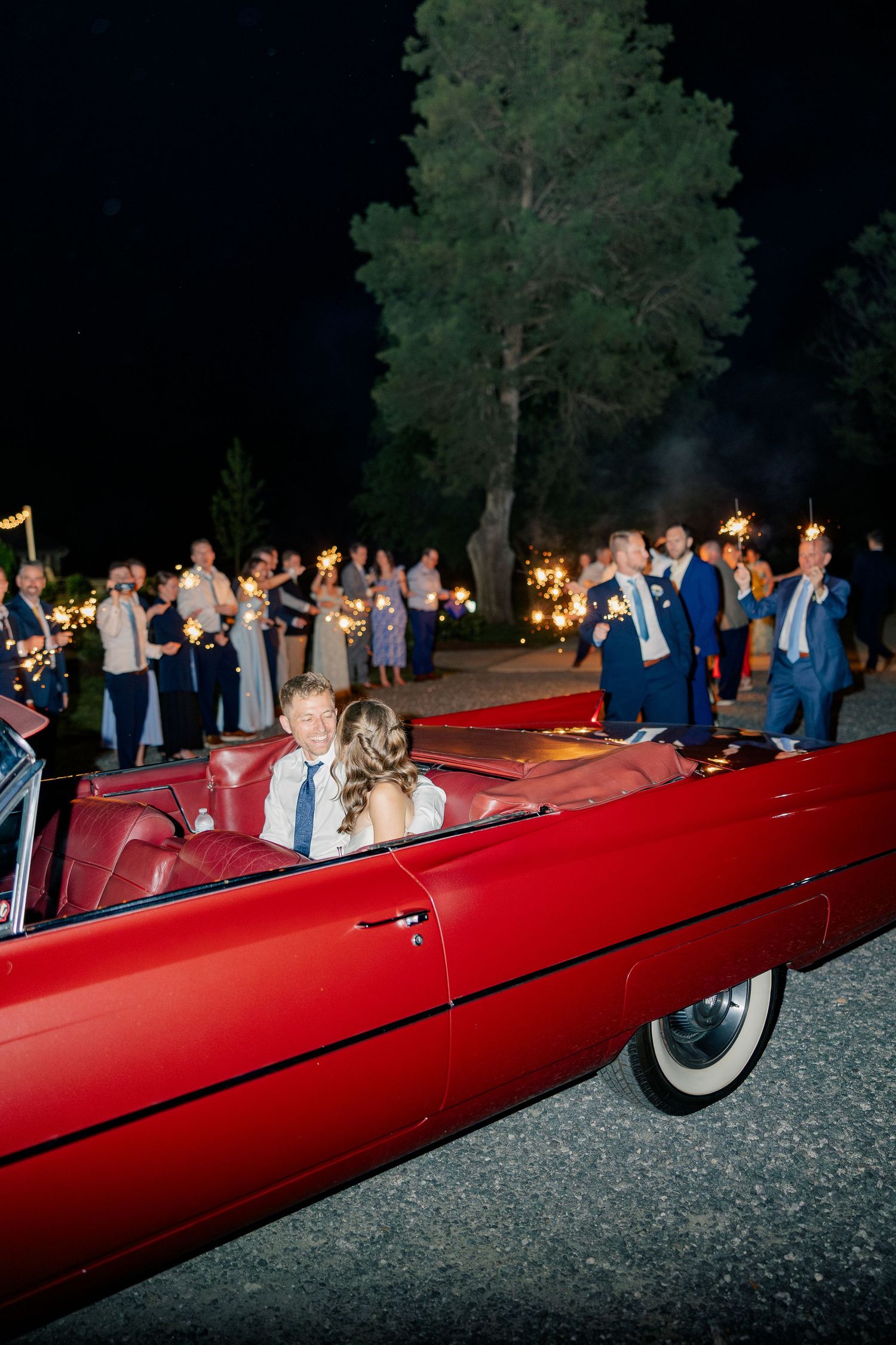 Newlyweds depart in vintage red convertible through sparkler send-off under night sky.