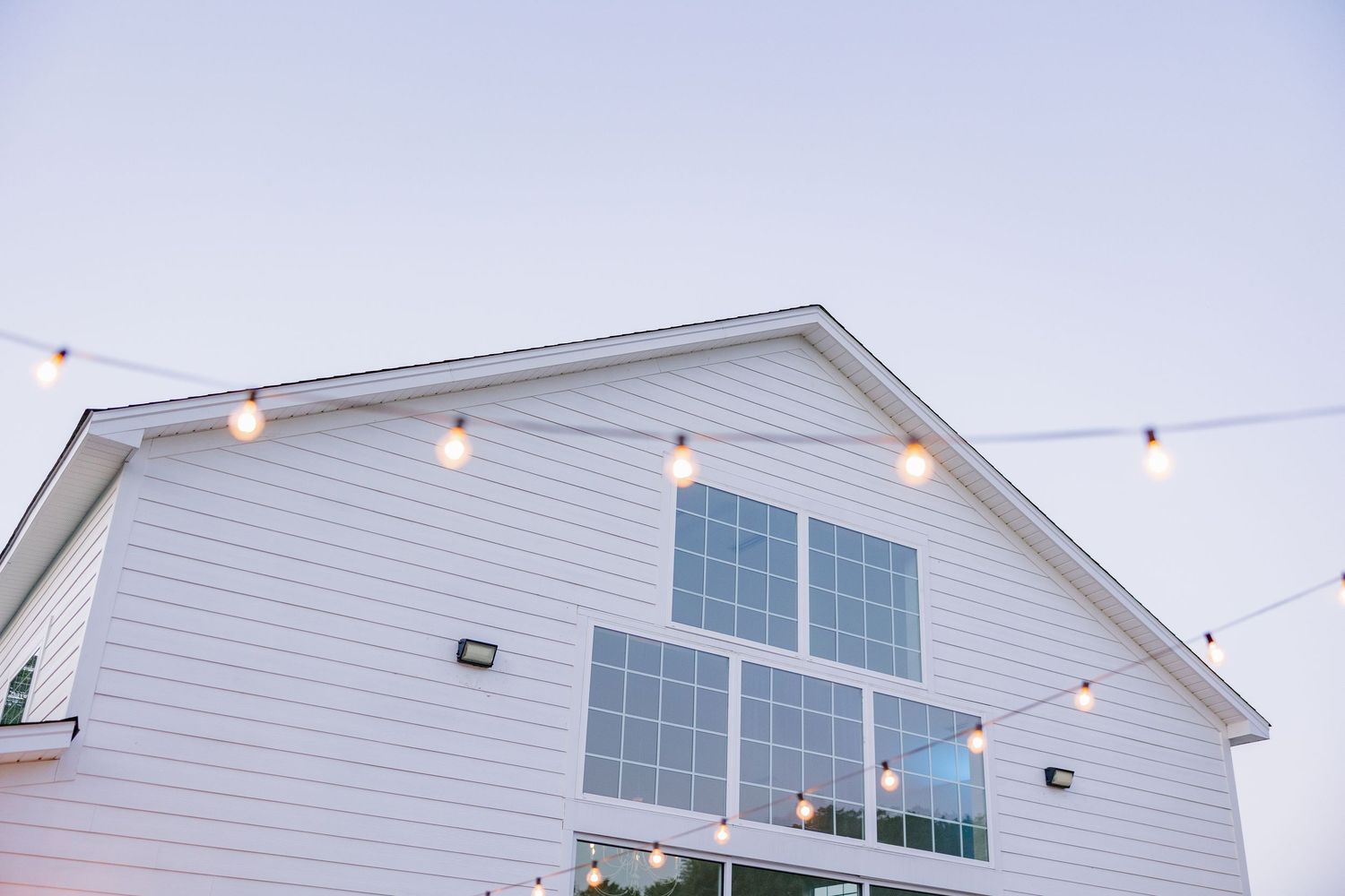 Elegant white barn wedding venue exterior with string lights against a soft blue twilight sky.