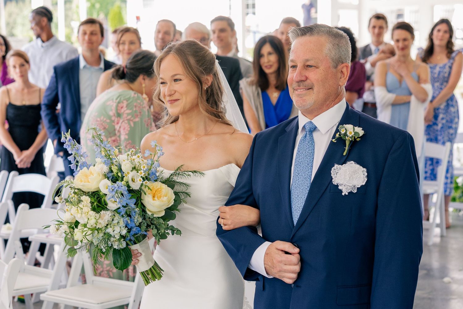 Bride walks down aisle with father holding blue and white bouquet at elegant wedding ceremony.