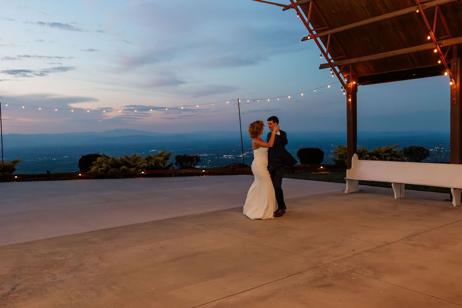 Bride and groom sharing first dance on outdoor patio under string lights at dusk.