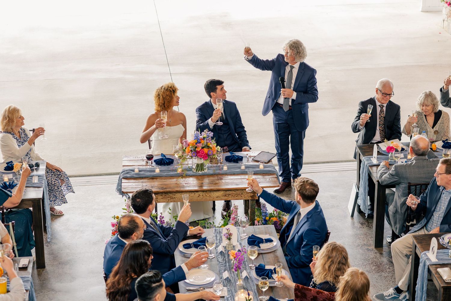 Wedding guests raising glasses during a toast at a reception with colorful floral centerpieces.