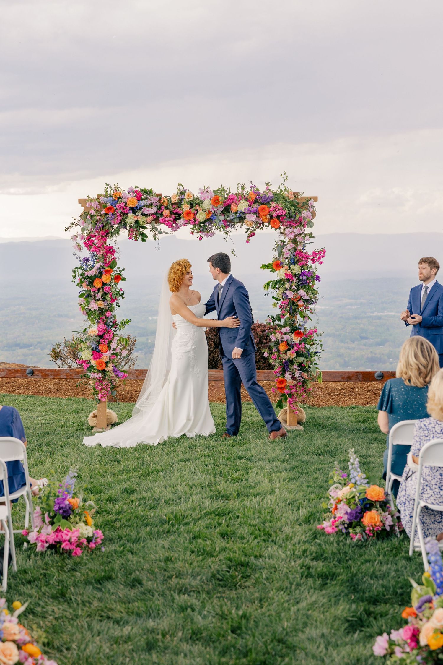 Bride and groom exchange vows under a colorful floral arch at an outdoor wedding ceremony with guests.