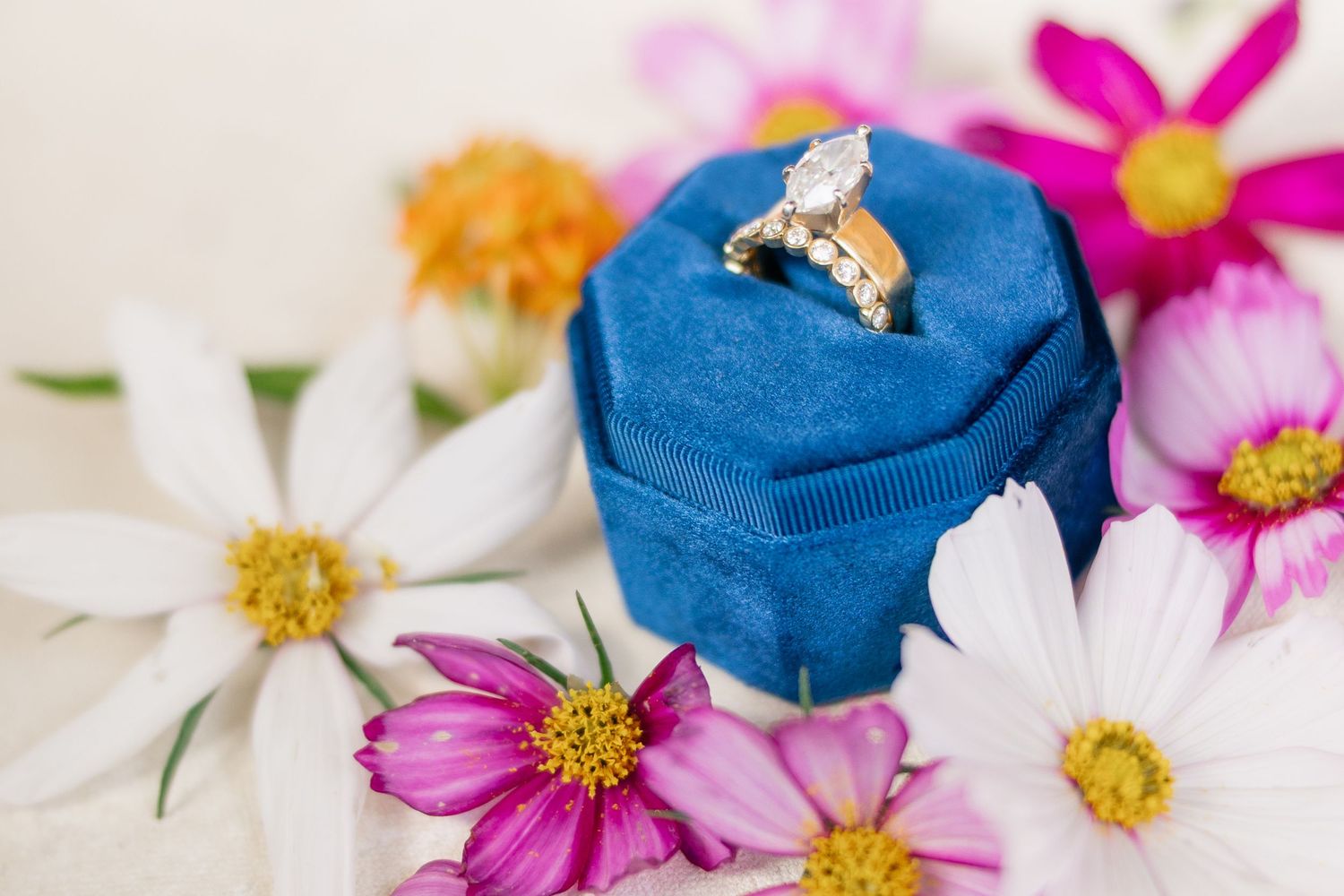 Gold diamond engagement ring displayed in a blue velvet box surrounded by colorful cosmos and daisy flowers.