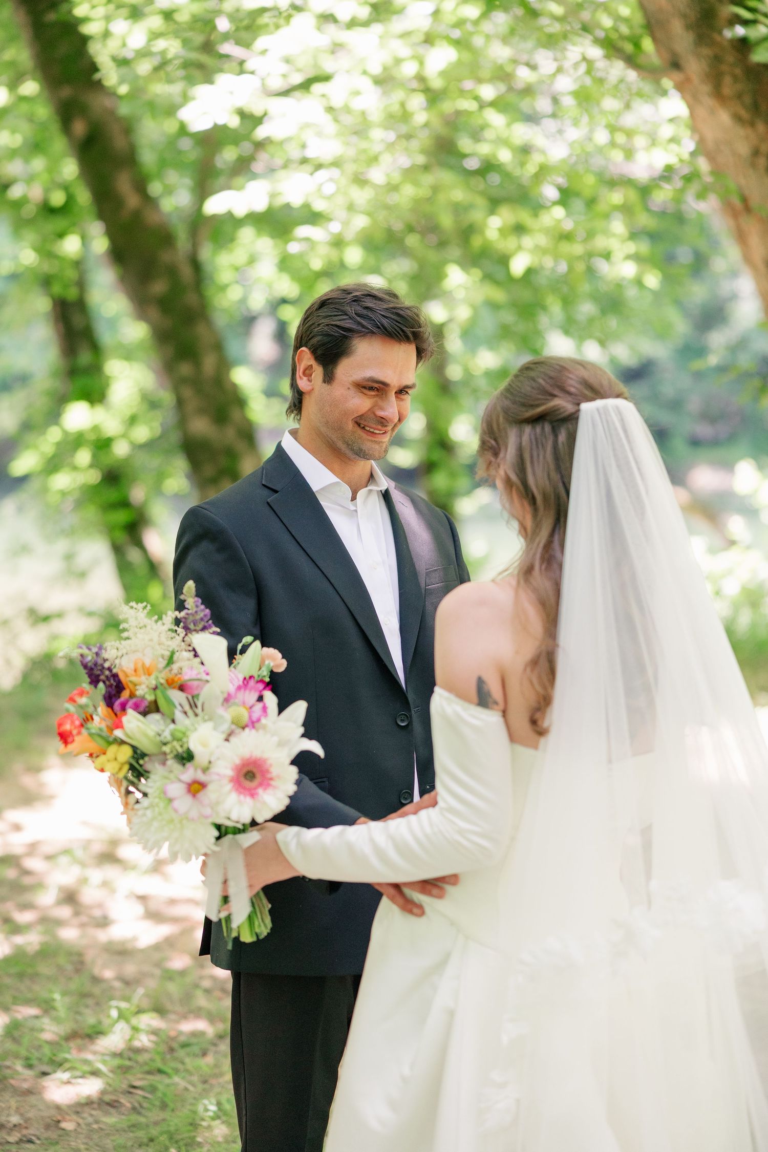 Smiling groom holds colorful bridal bouquet while facing bride in white dress and veil outdoors.