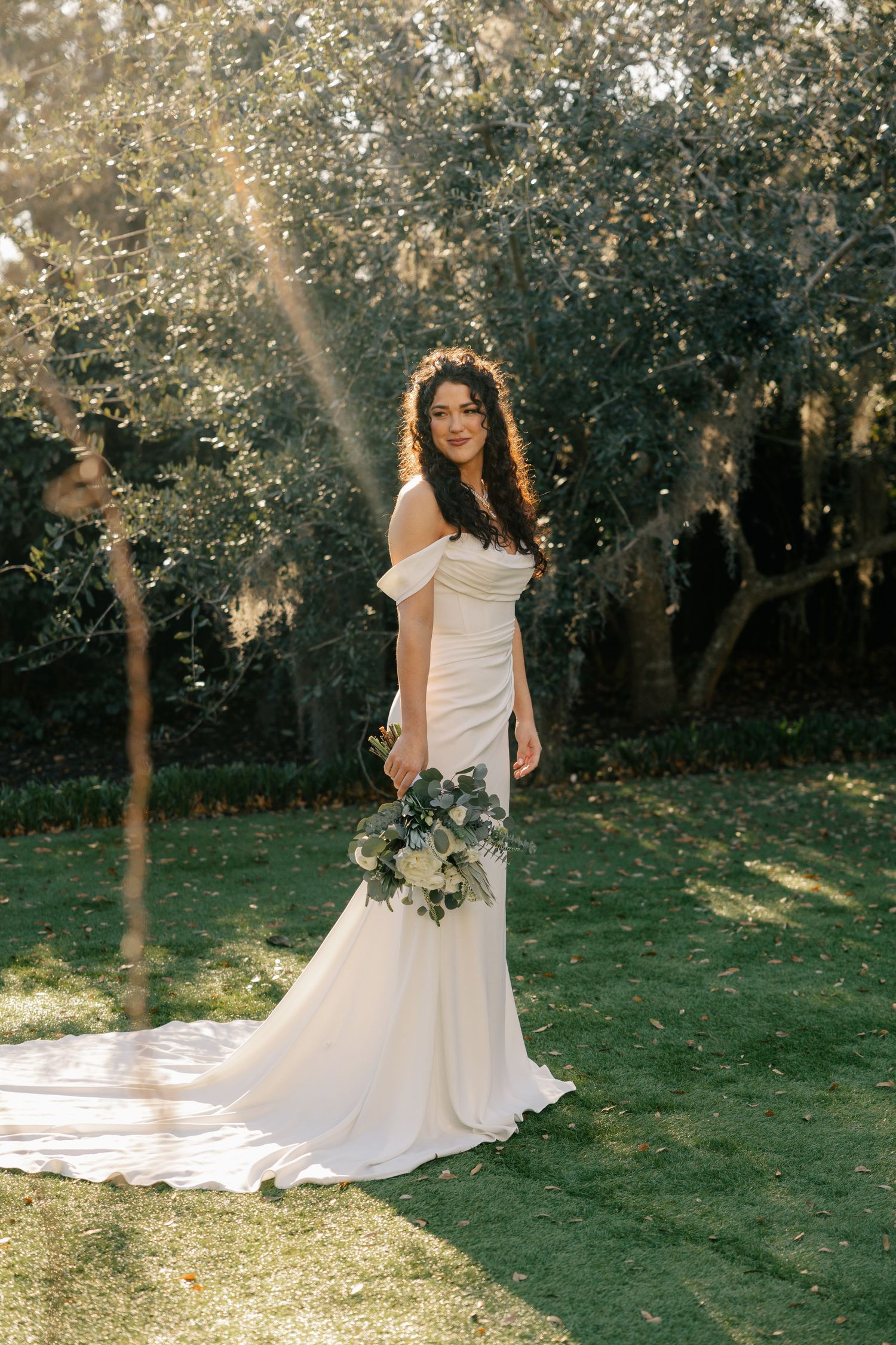 A white wedding gown catches golden sunlight in an outdoor garden setting with trees in the background.