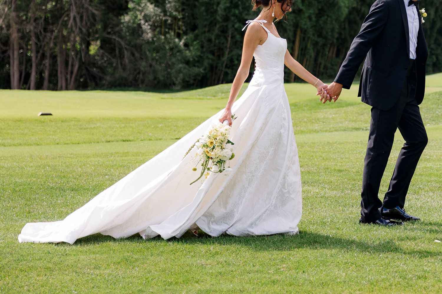 Bride in a white lace gown holds a bouquet while walking hand-in-hand with groom on a lush green lawn.