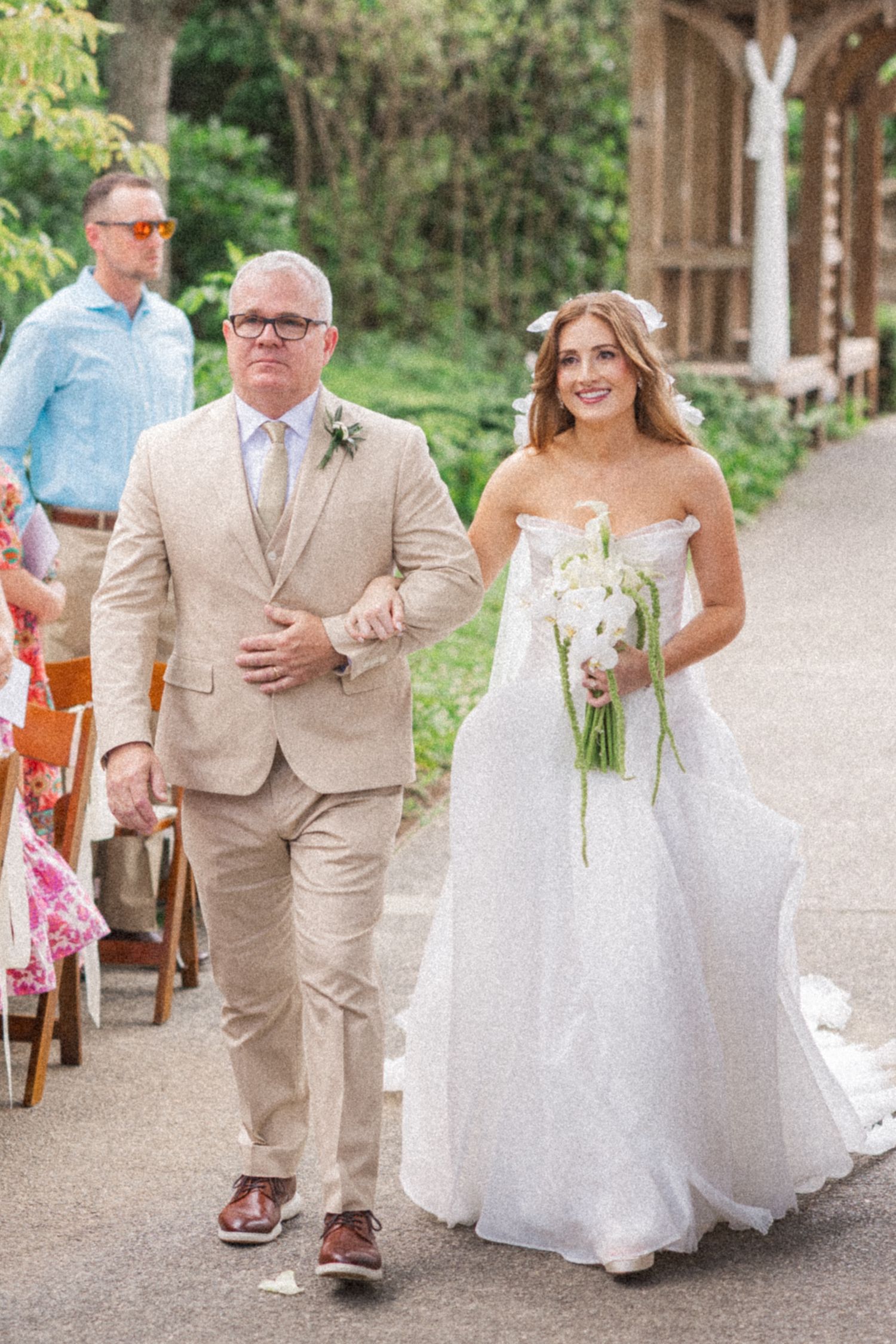 A wedding couple walks down a tree-lined path in matching beige and white attire during an outdoor ceremony.