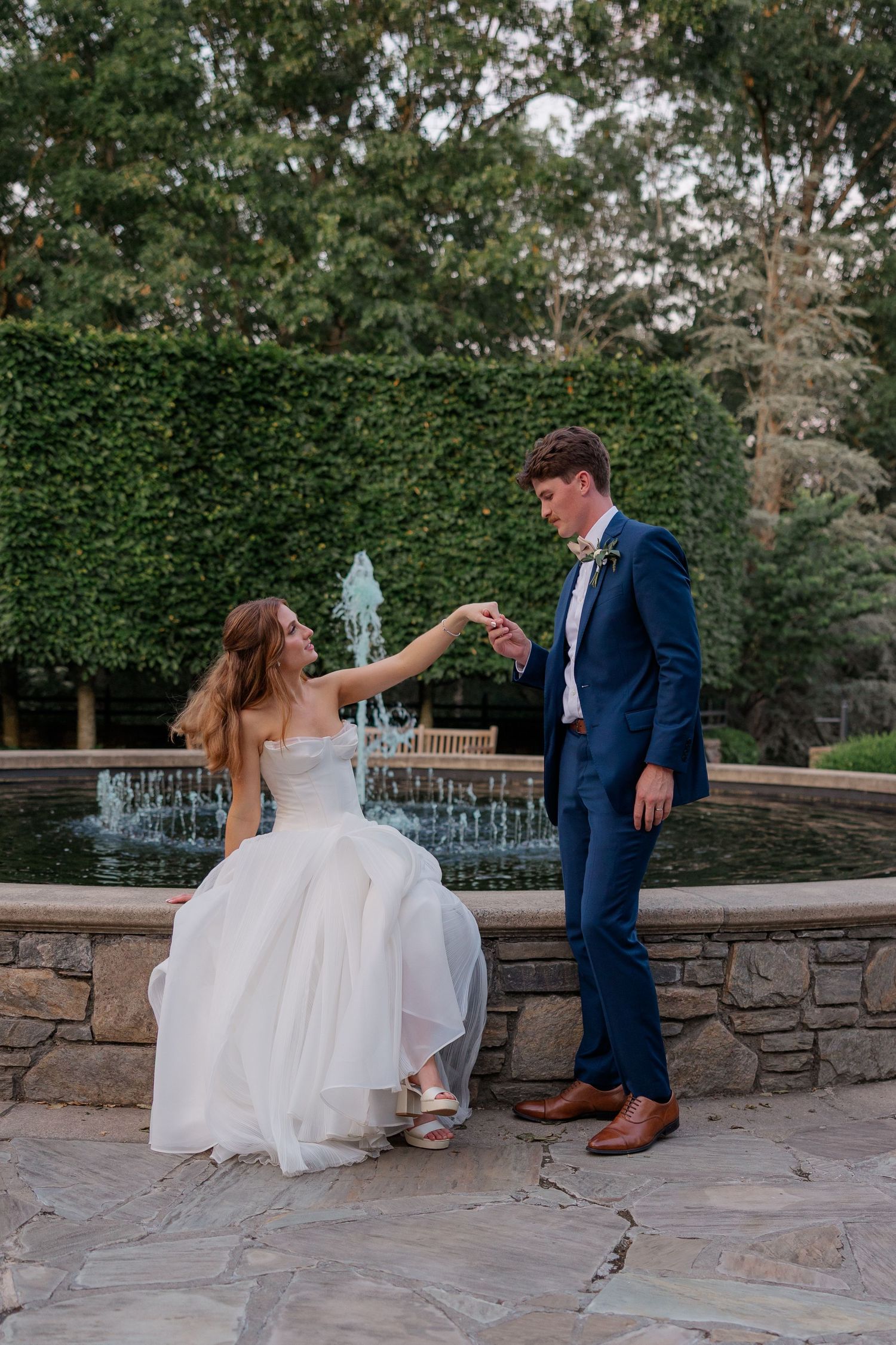 Bride and groom share a playful moment on stone patio near garden fountain during wedding photo session.