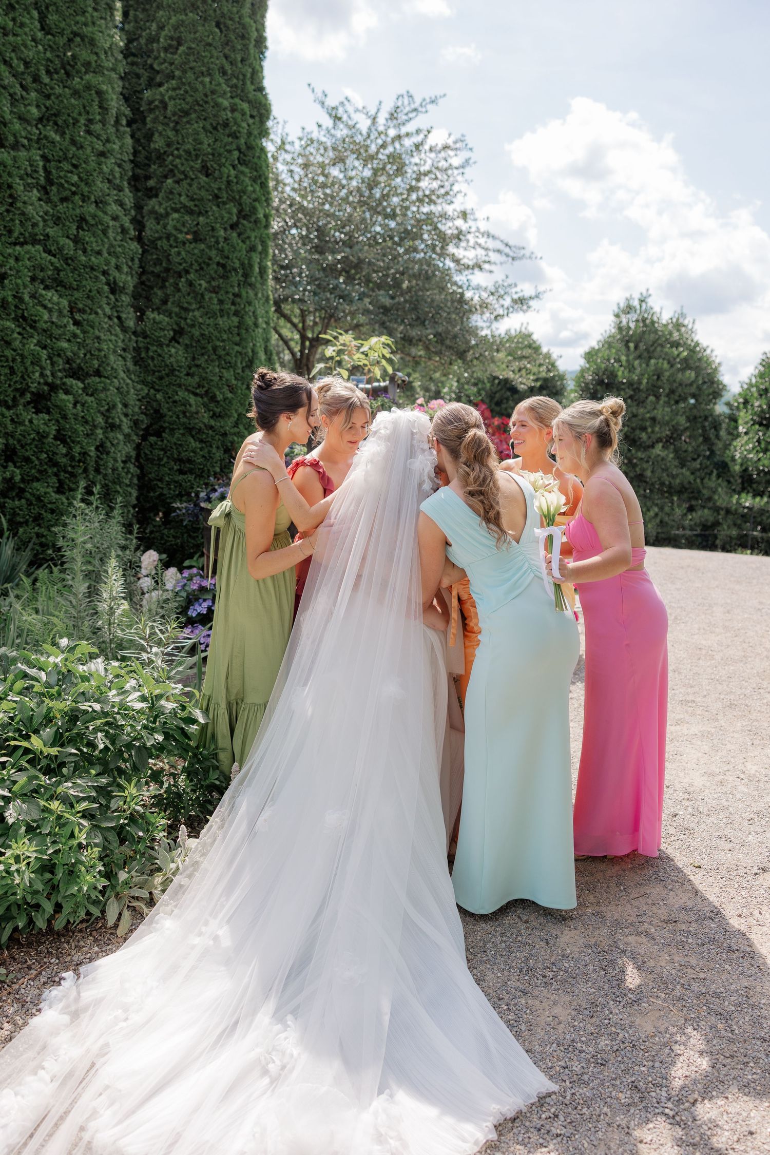 A group in pastel colored dresses help adjust a flowing white wedding gown train in an outdoor garden setting.