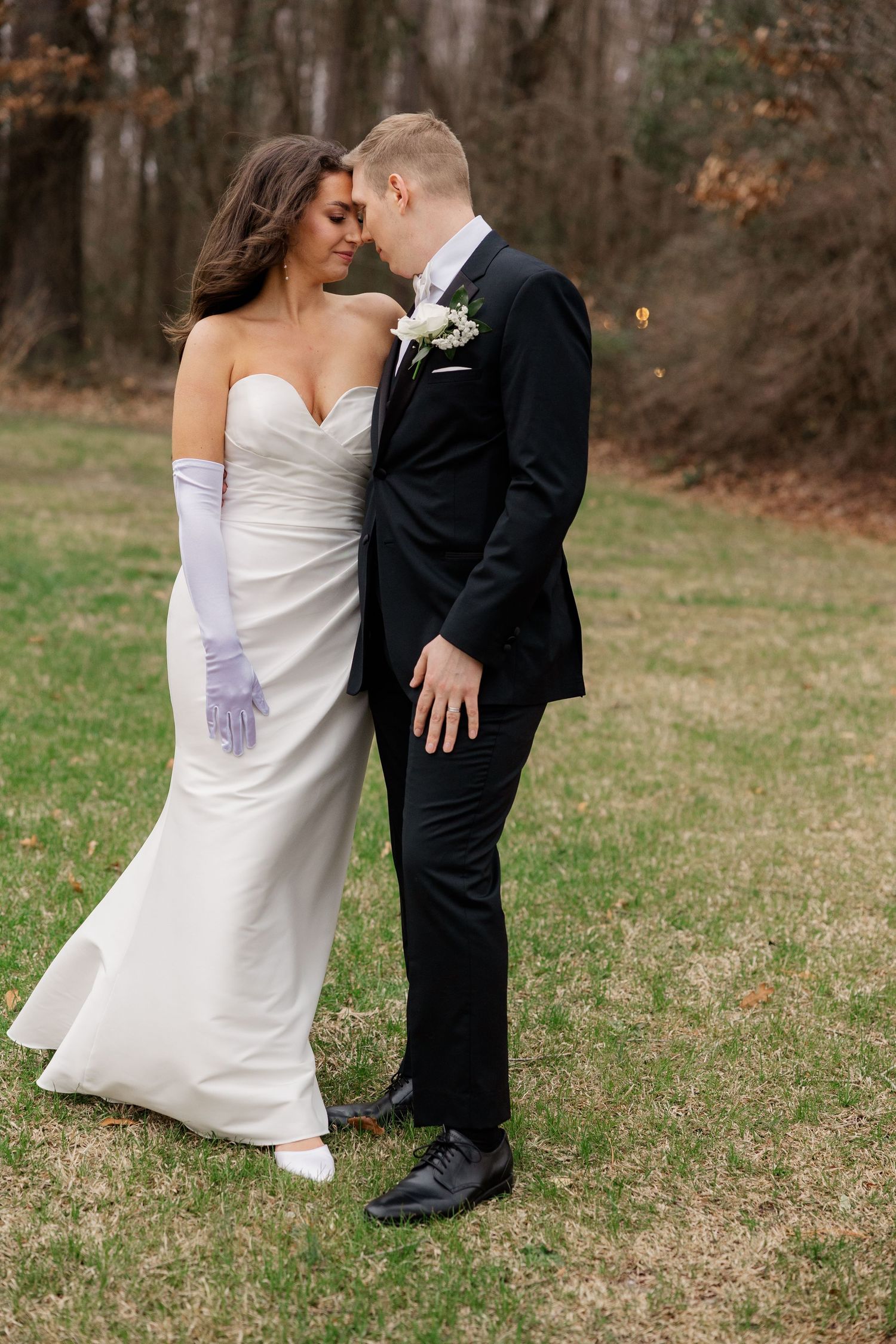 An elegant wedding couple shares an intimate moment on a grassy lawn with trees in the background.