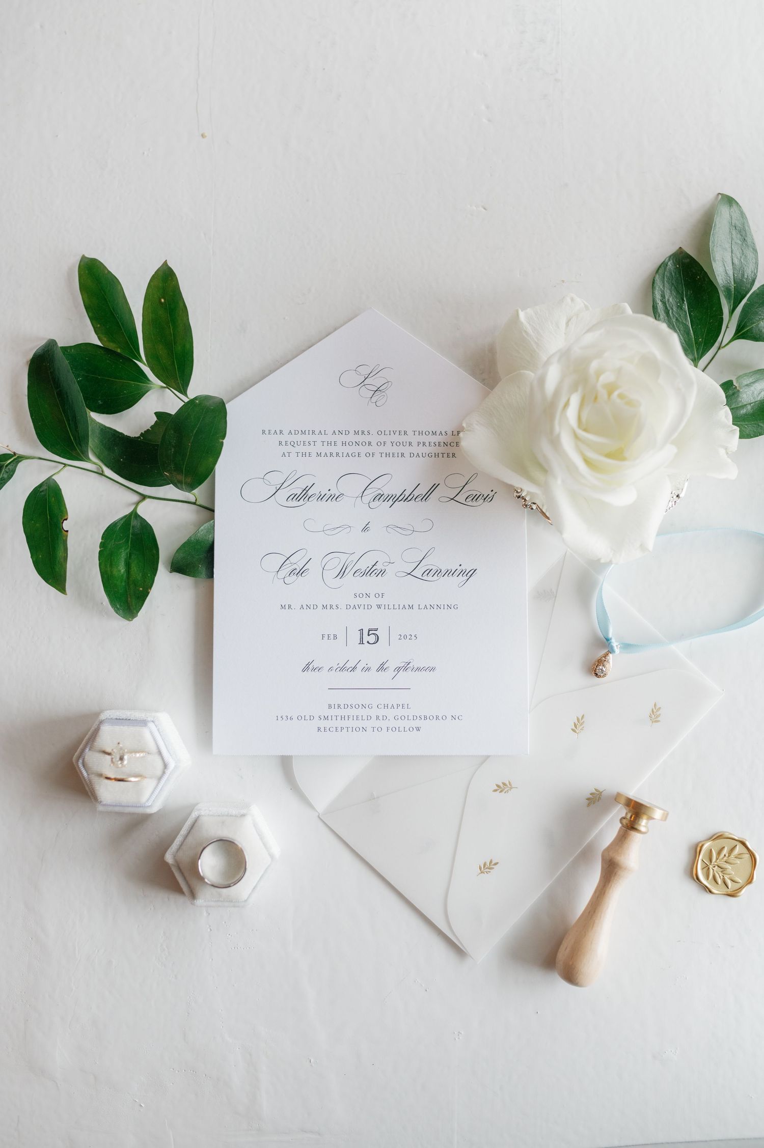 Wedding stationery set with eucalyptus leaves, white roses, and gold wax seals on marble surface.