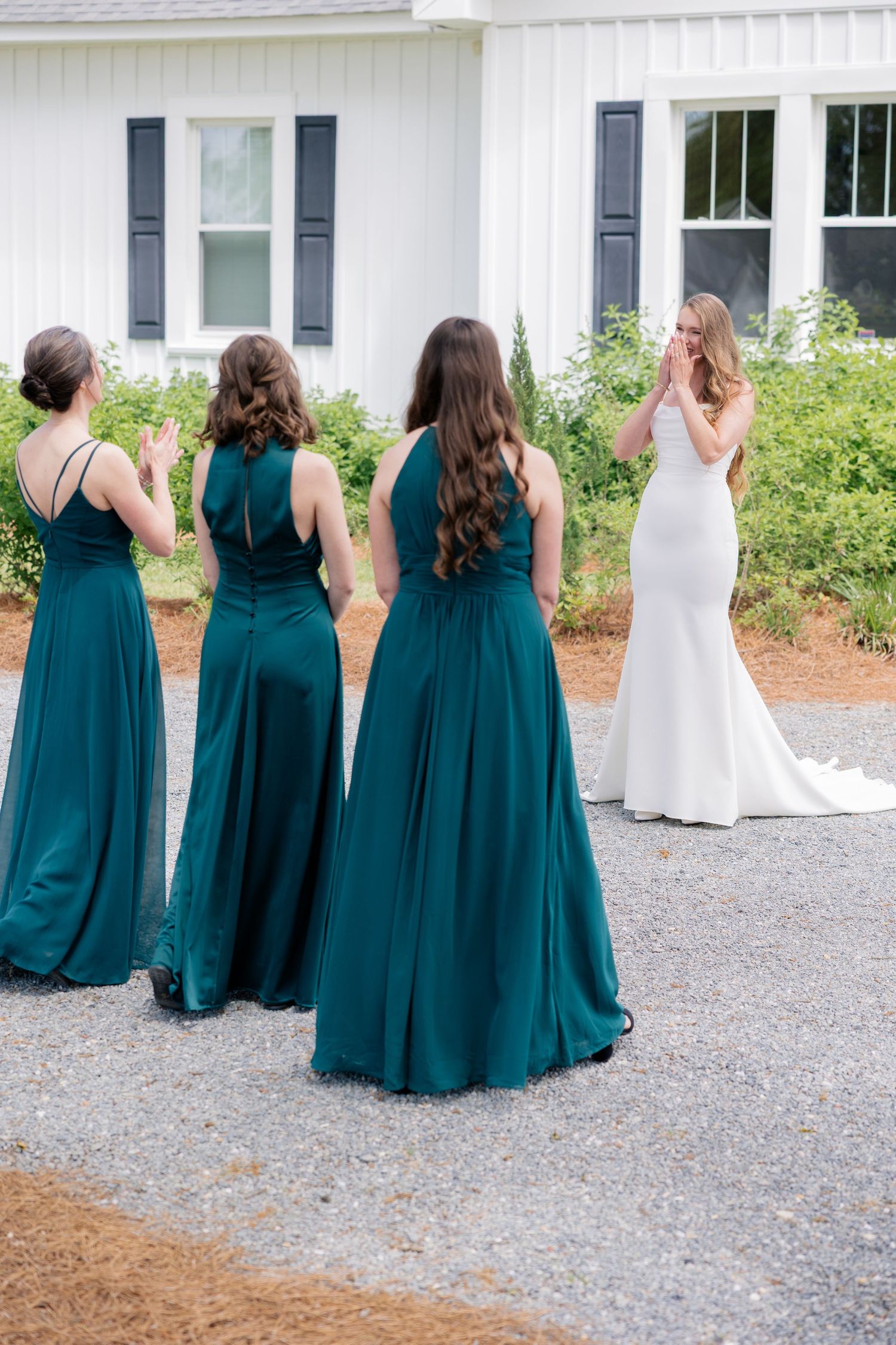 Bridesmaids in elegant teal full-length dresses standing behind bride wearing white wedding gown on gravel path.