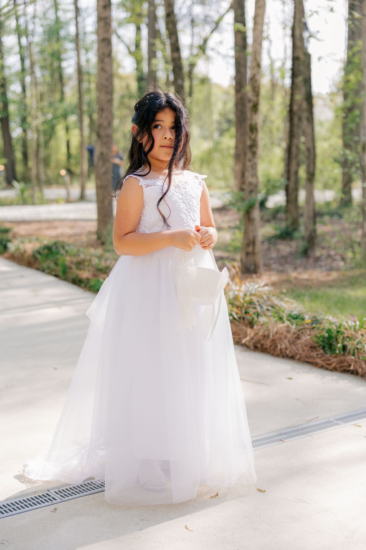 Bride in flowing white gown poses along a paved pathway in a natural forest setting.