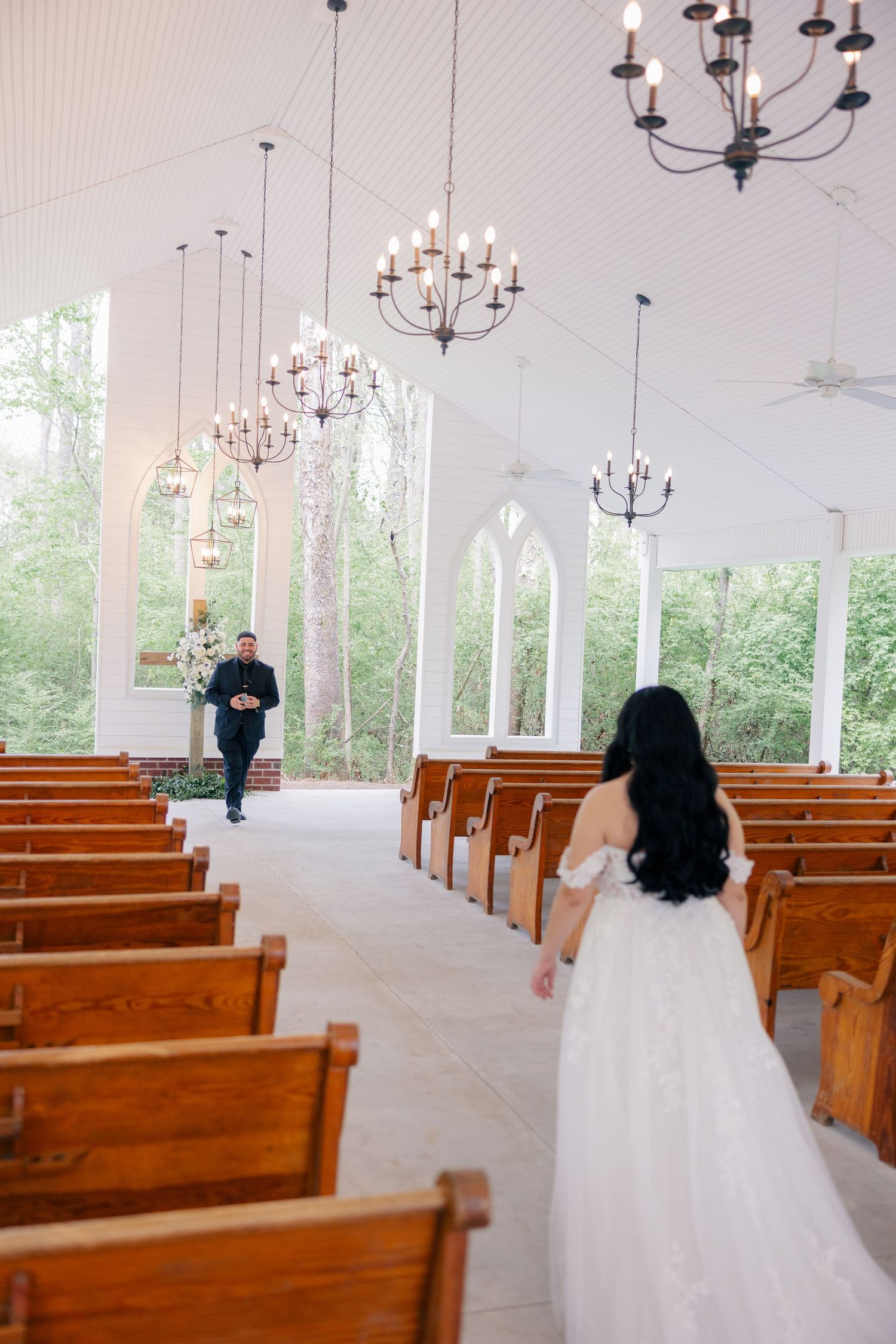 A spacious chapel with wooden pews and chandeliers features white walls and large windows.