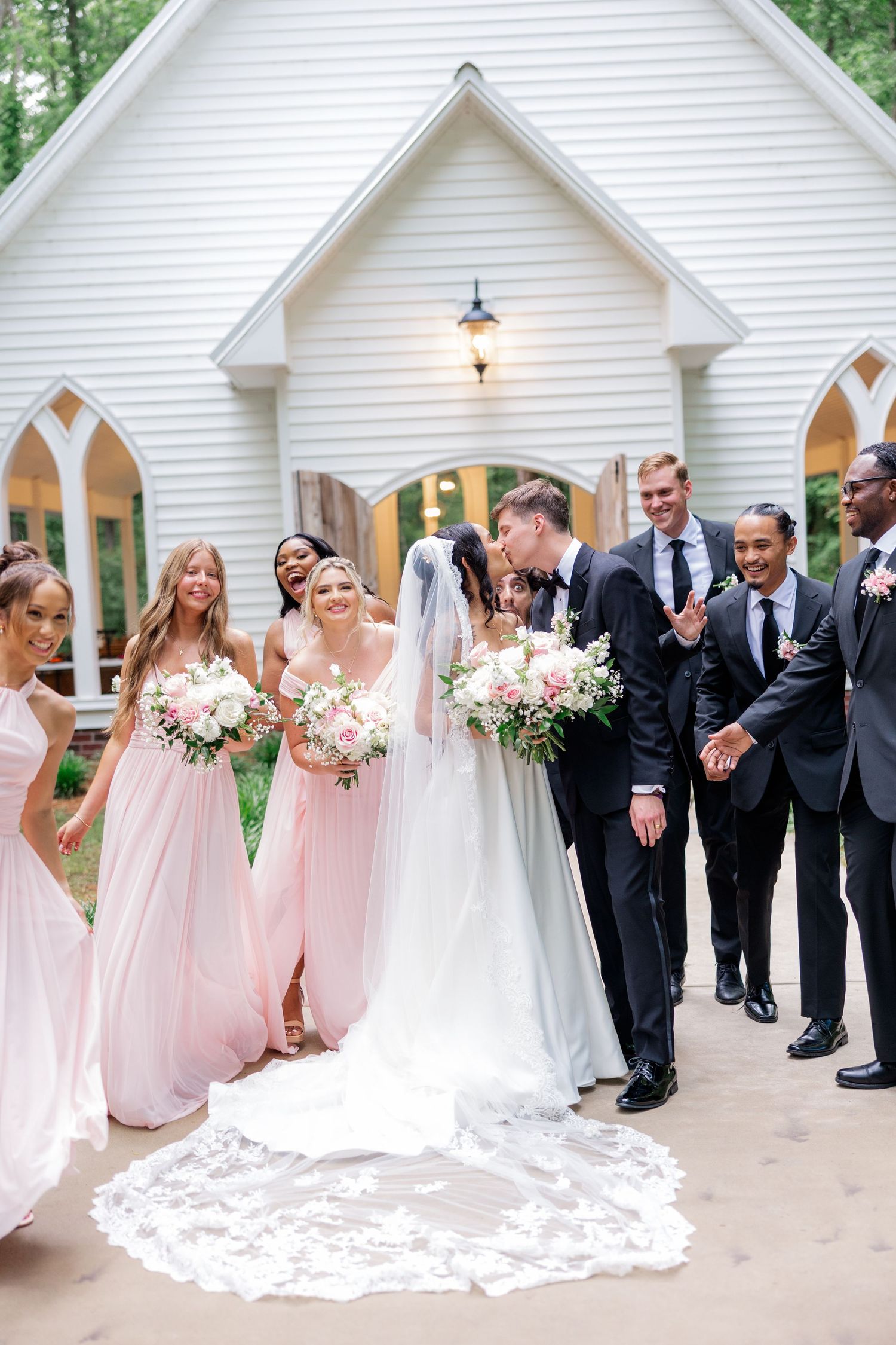 Wedding party poses outside white chapel with bridesmaids in pink gowns.