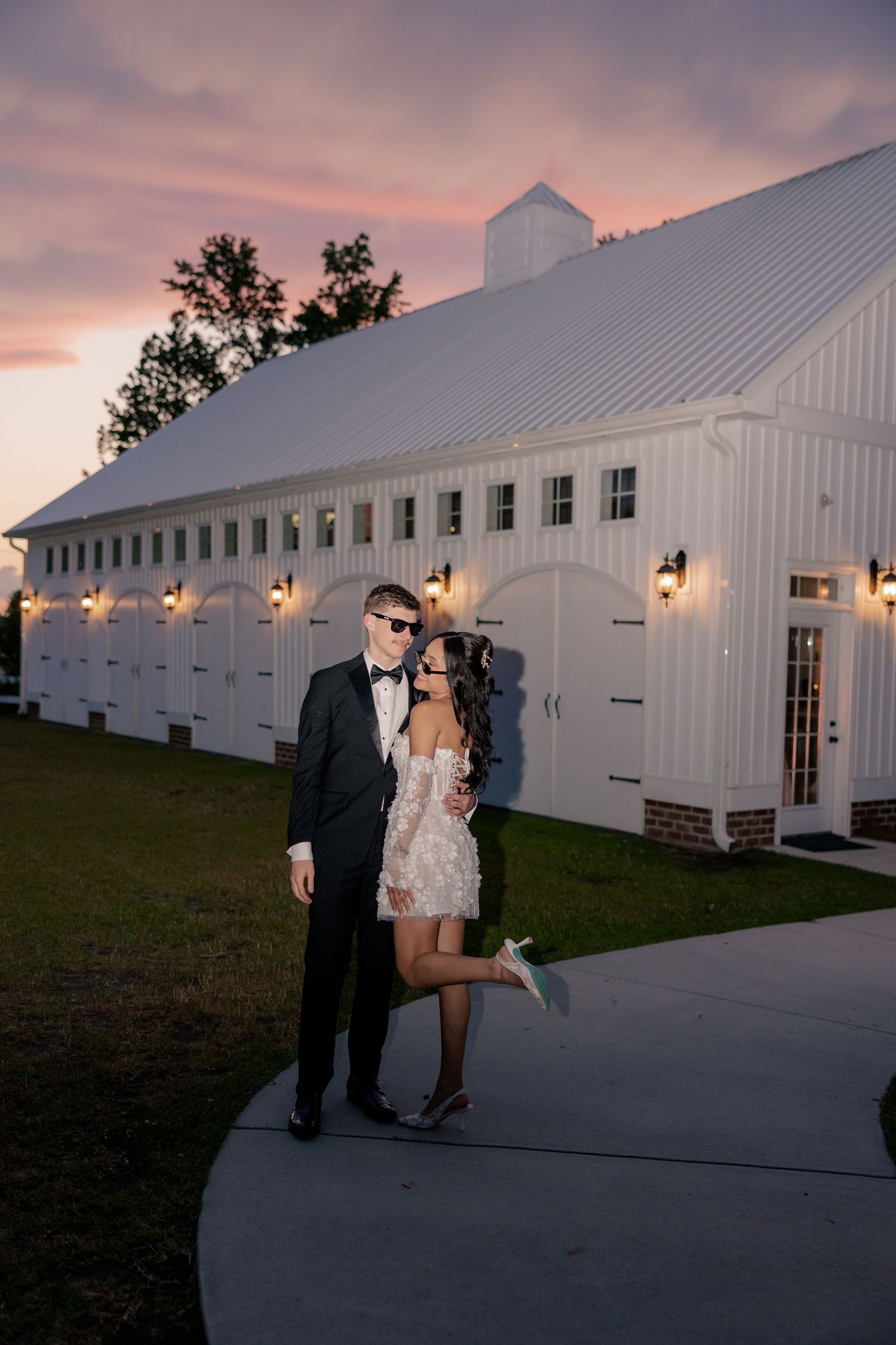 Wedding couple poses outside a white barn venue at sunset with wall-mounted lanterns.