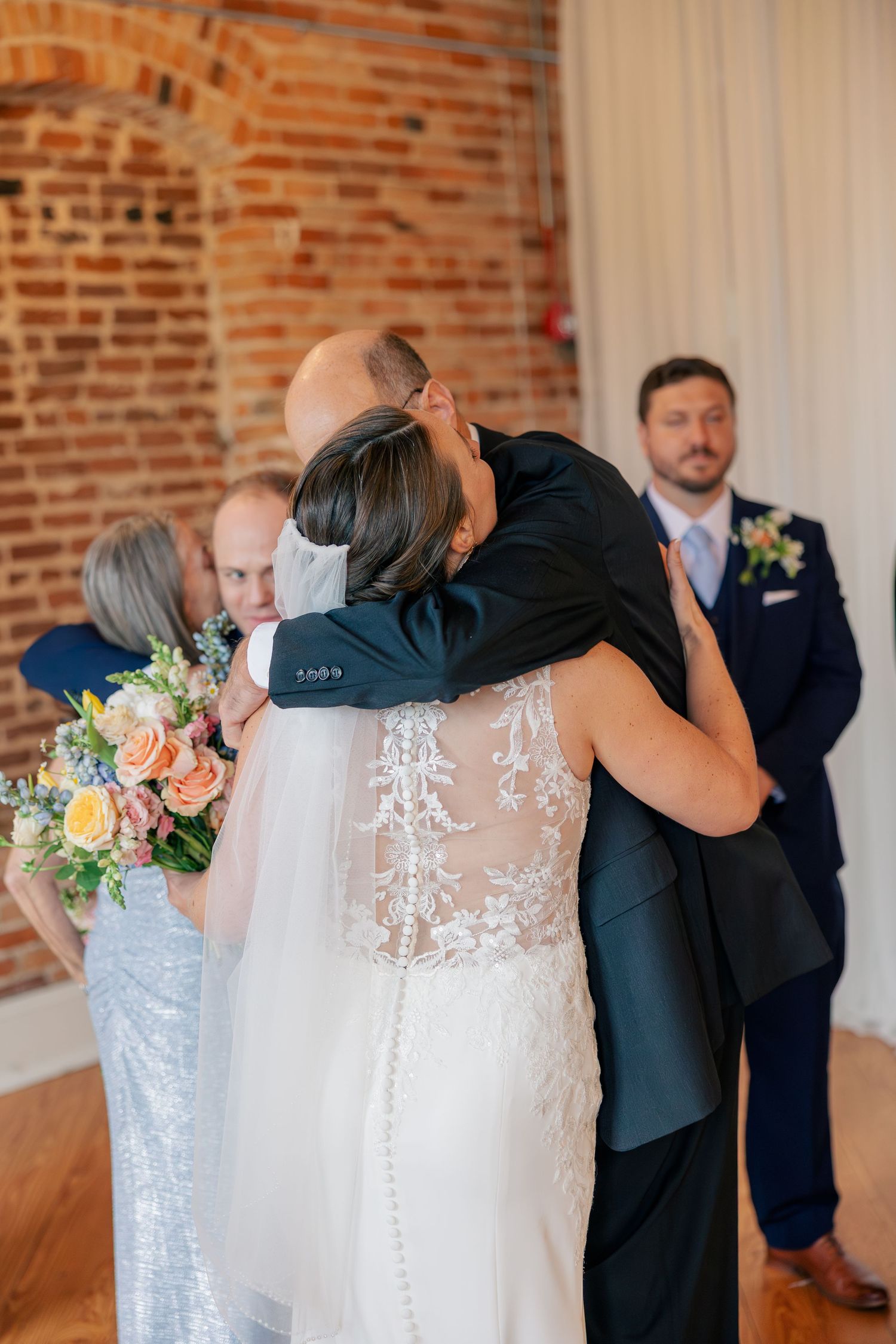 Bride in lace wedding gown embraces family member in emotional hug at rustic brick venue.