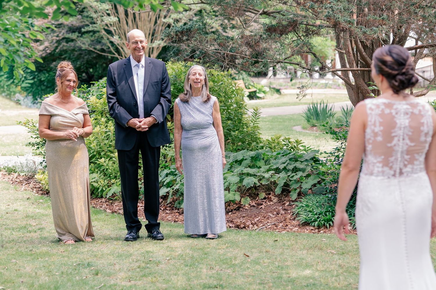 Bride in lace wedding gown seen from behind, facing smiling family members in formal attire outdoors in a lush garden.