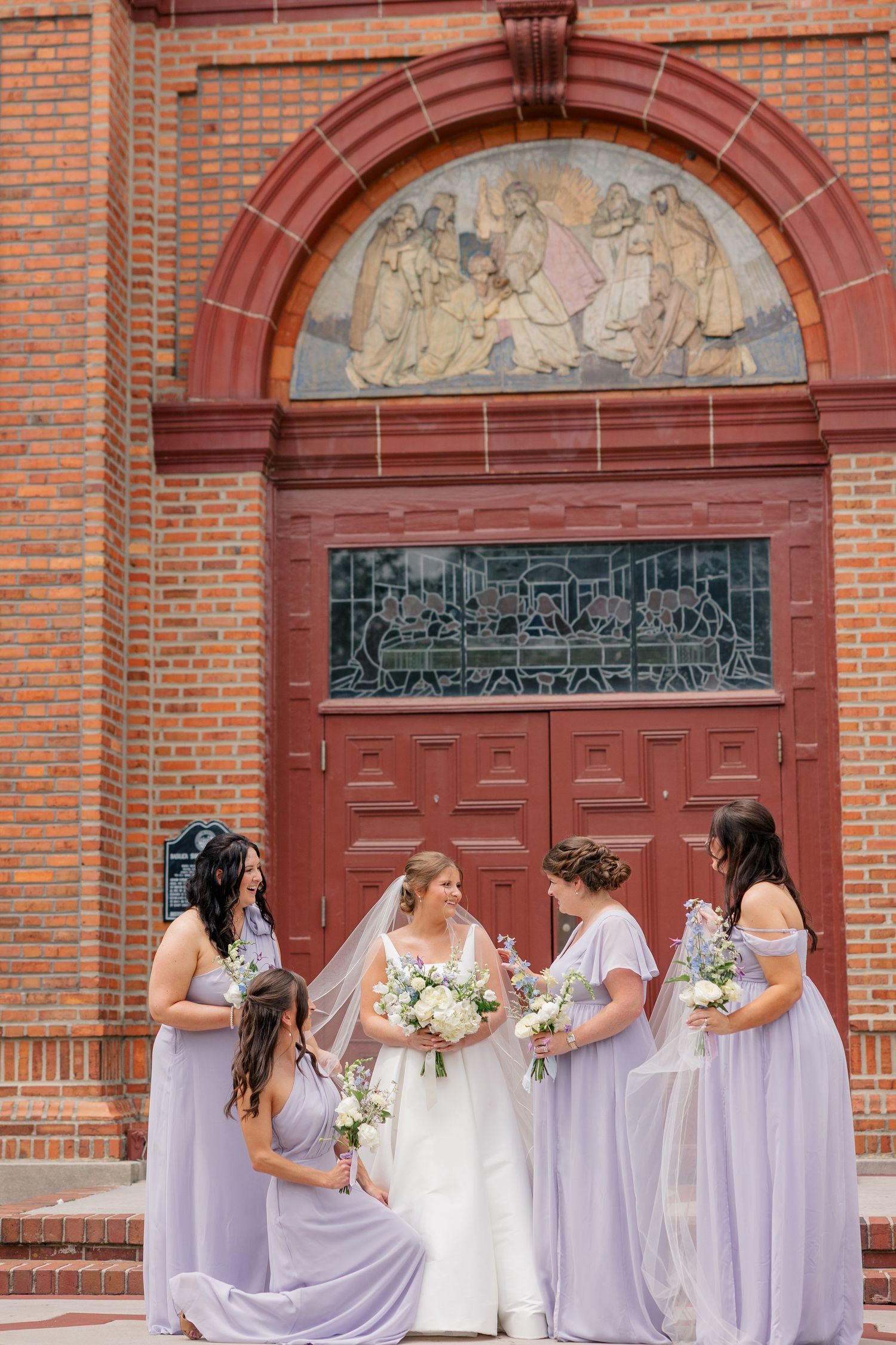 Bride with bridesmaids in lavender gowns pose before ornate red brick church entrance doors.