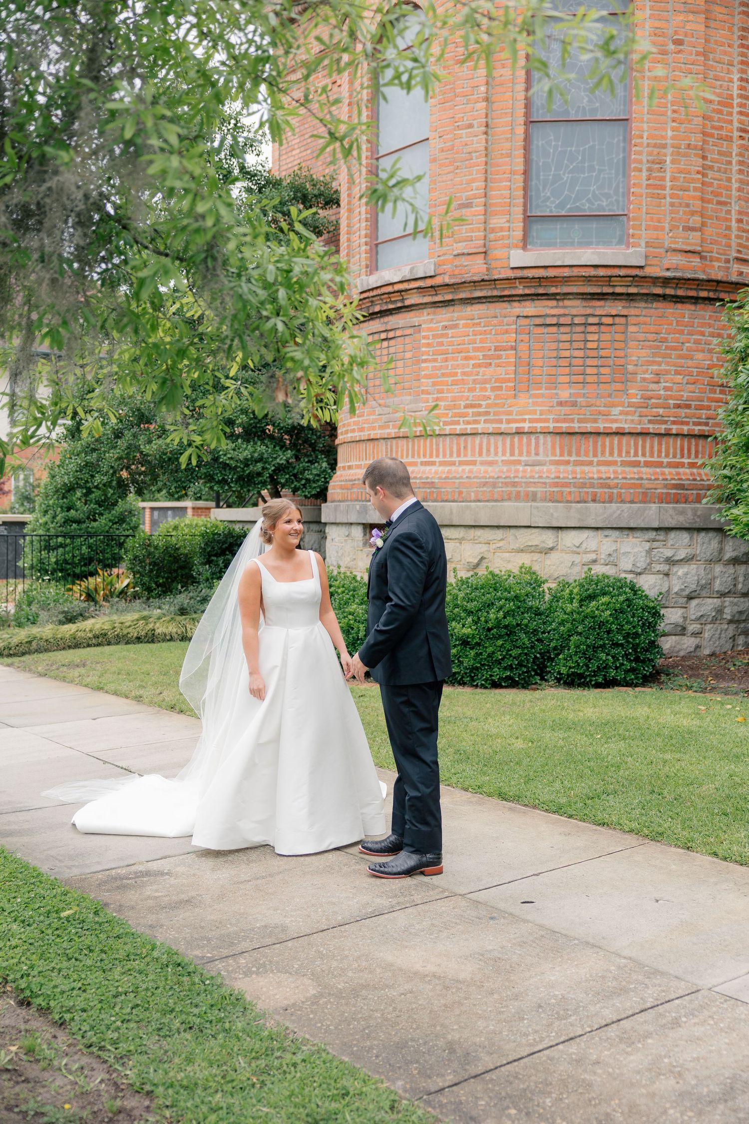 Bride in flowing white gown with veil holds hands with groom outside historic brick church on wedding day.