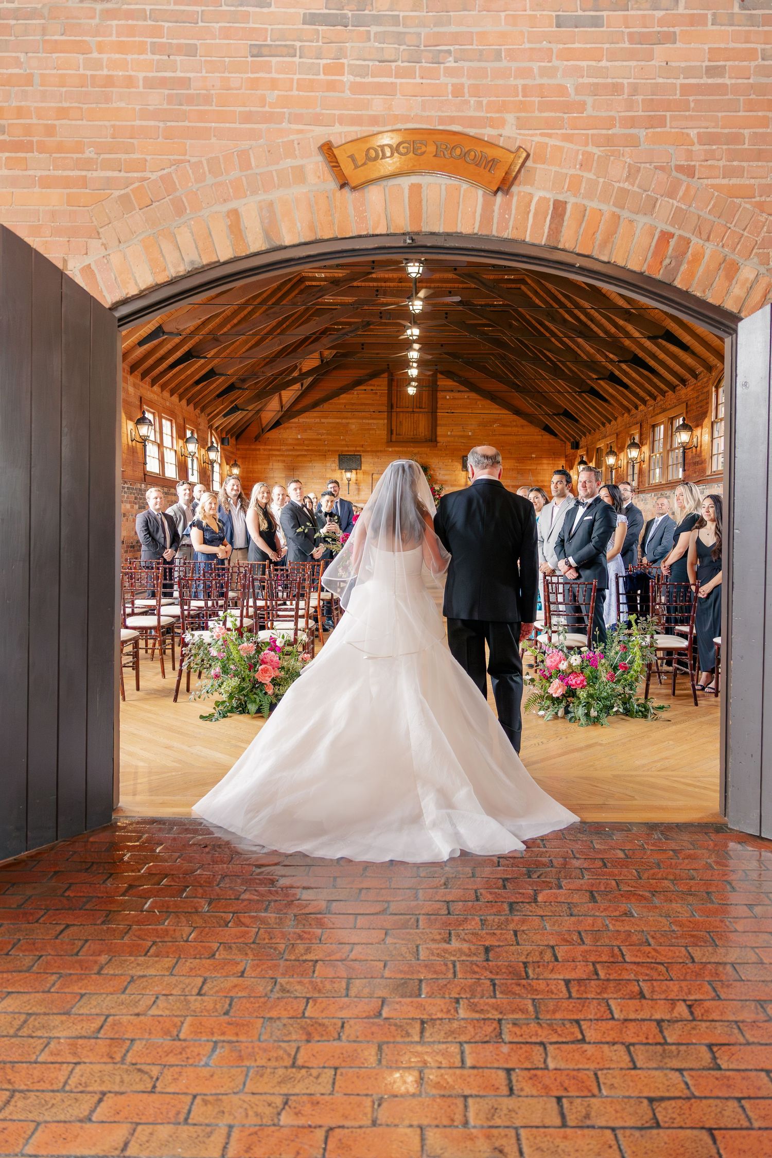 Bride and father walk through brick archway into elegant wood-beamed wedding ceremony venue filled with guests.