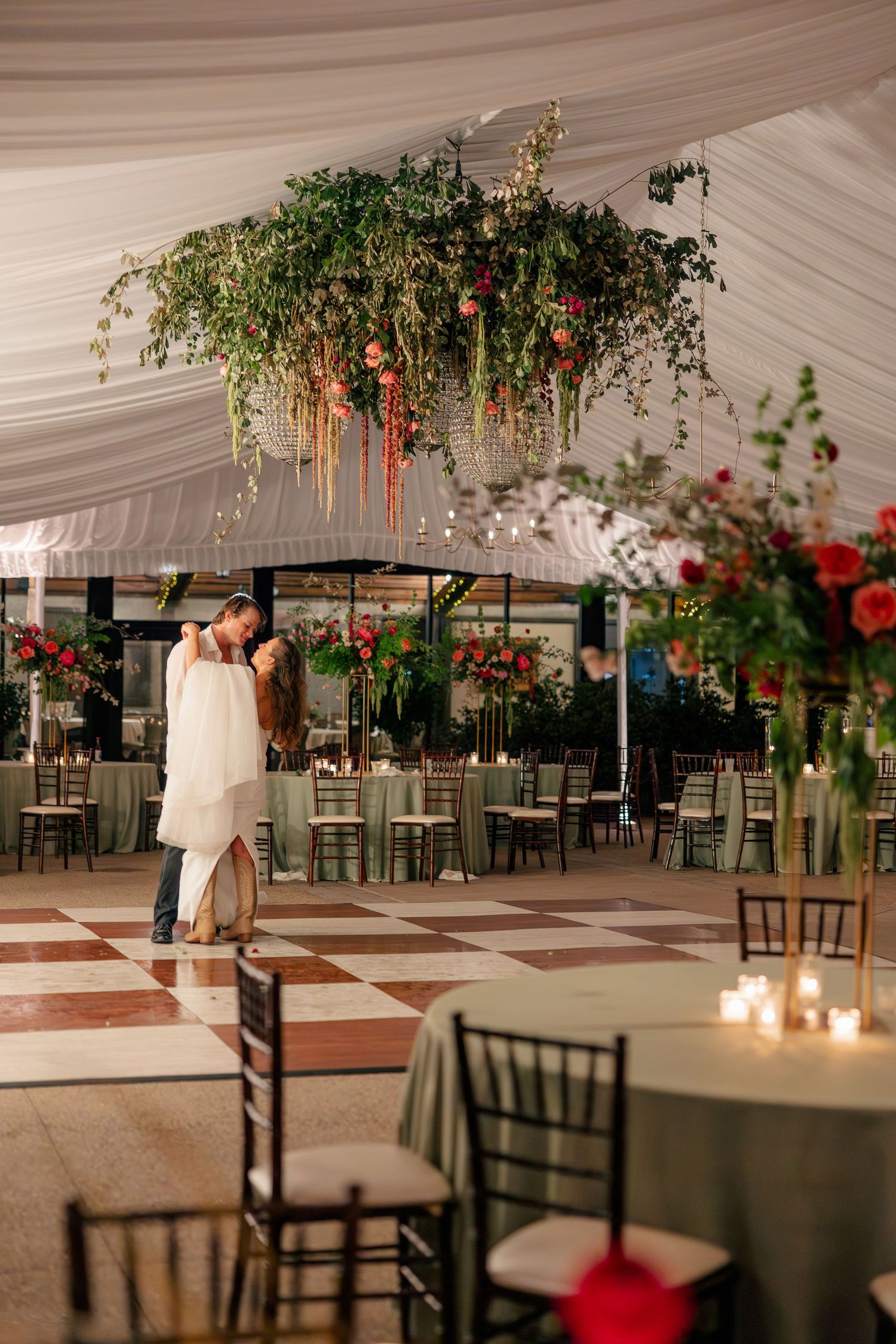 Couple shares last dance in empty tent venue under lush floral installation on checkered dance floor.