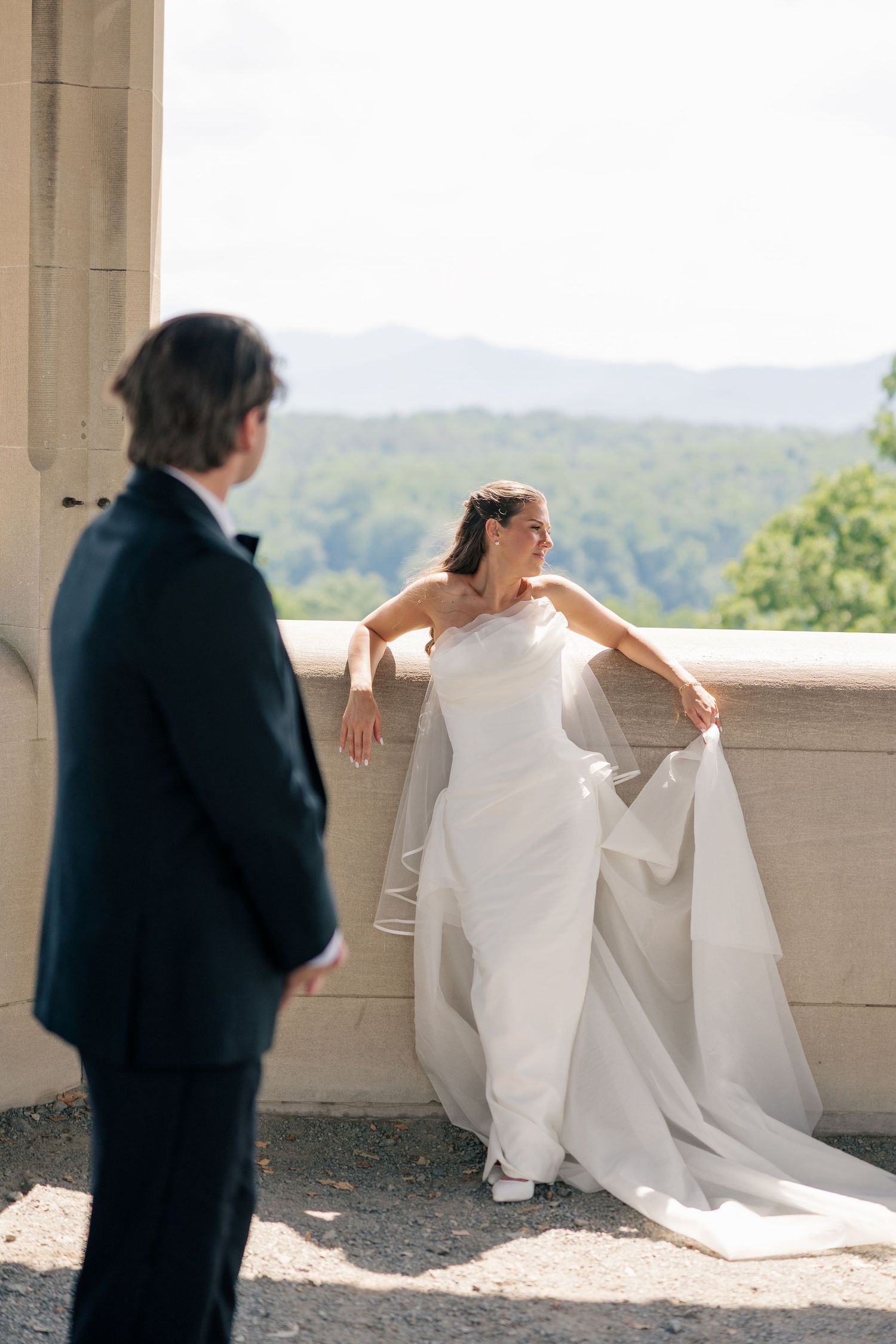 Bride in elegant one-shoulder gown poses on balcony with mountain views and groom nearby.