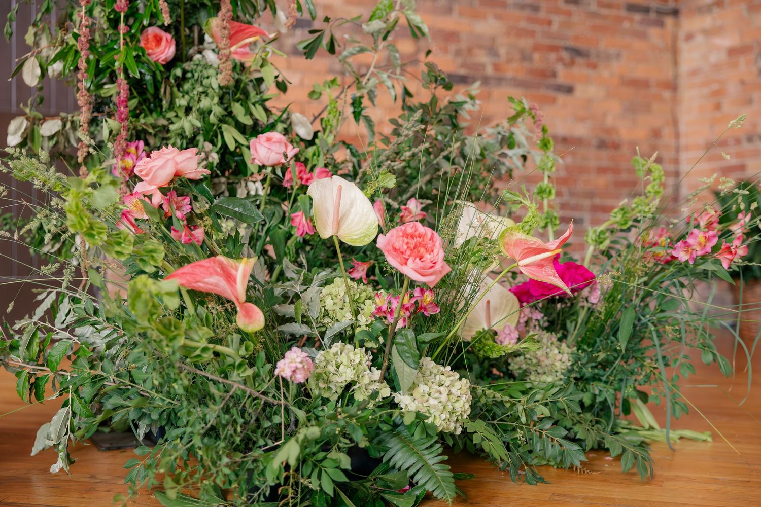Lush floral arrangement featuring pink anthuriums, roses, and greenery against a rustic brick backdrop.