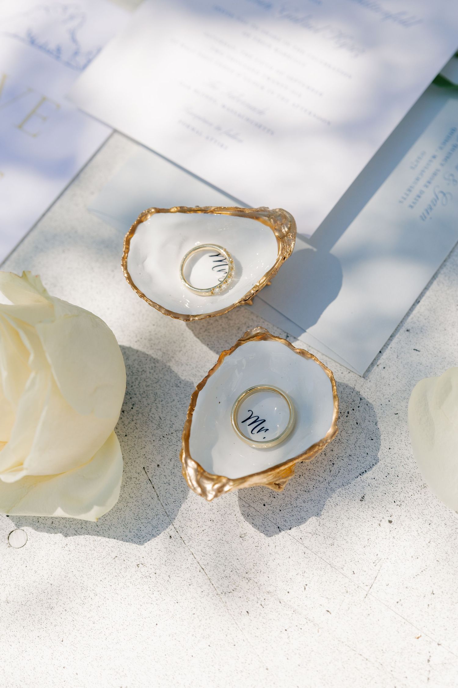 Gold-rimmed oyster shell ring dishes holding wedding bands on white surface with roses and invitation.